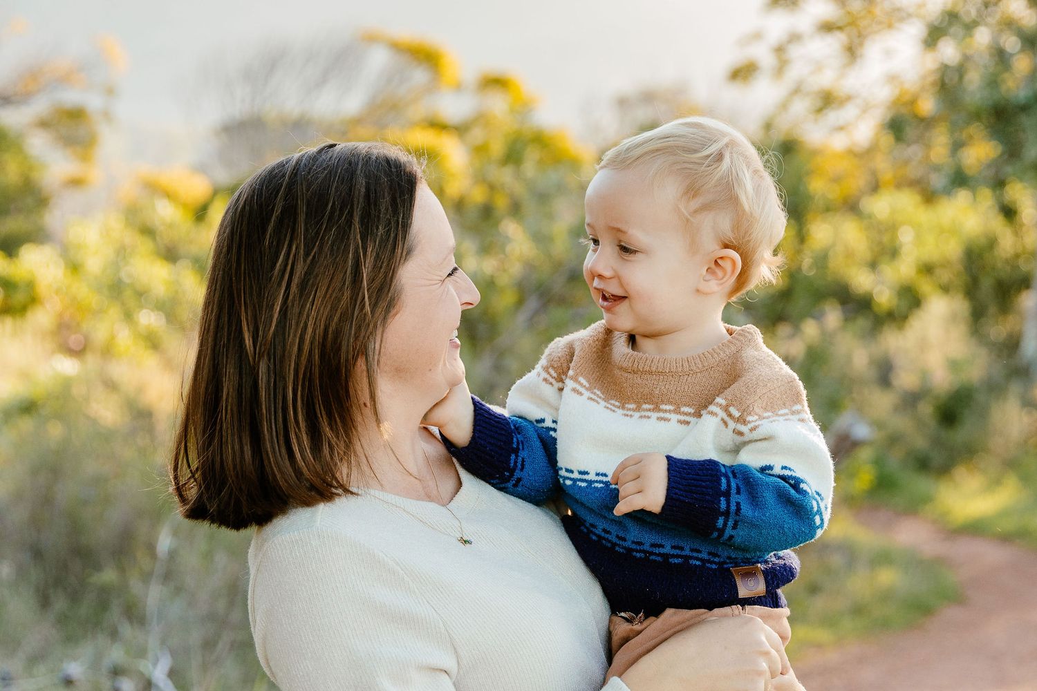 Mother smiling at cheerful blonde toddler she holds in golden outdoor setting.