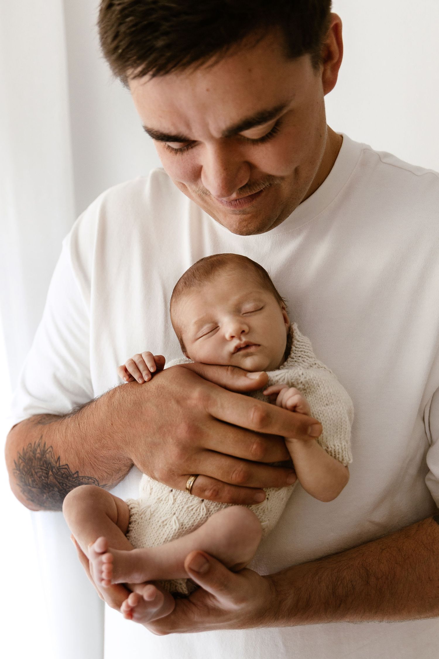 Proud father in white t-shirt smiles while holding sleeping newborn baby.