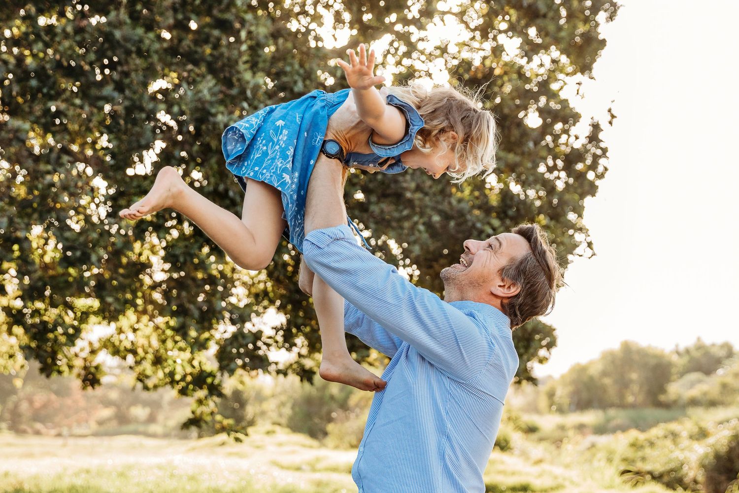 A joyful father lifts his smiling young daughter in a blue dress high into the air outdoors near a large tree.