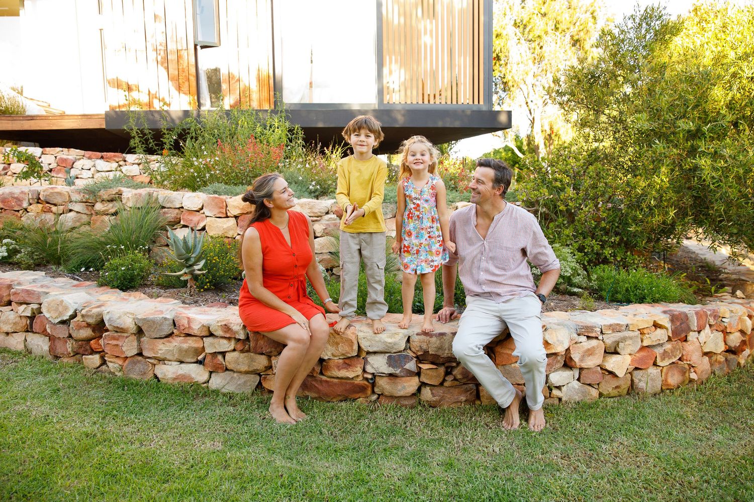 Family of four laughing together on a stone garden wall beside a modern tiny home surrounded by lush greenery.