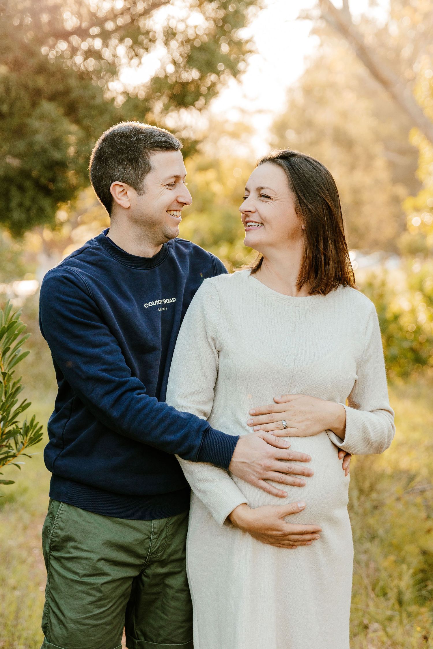 Happy couple embracing outdoors, man in navy sweatshirt with pregnant woman in white dress.