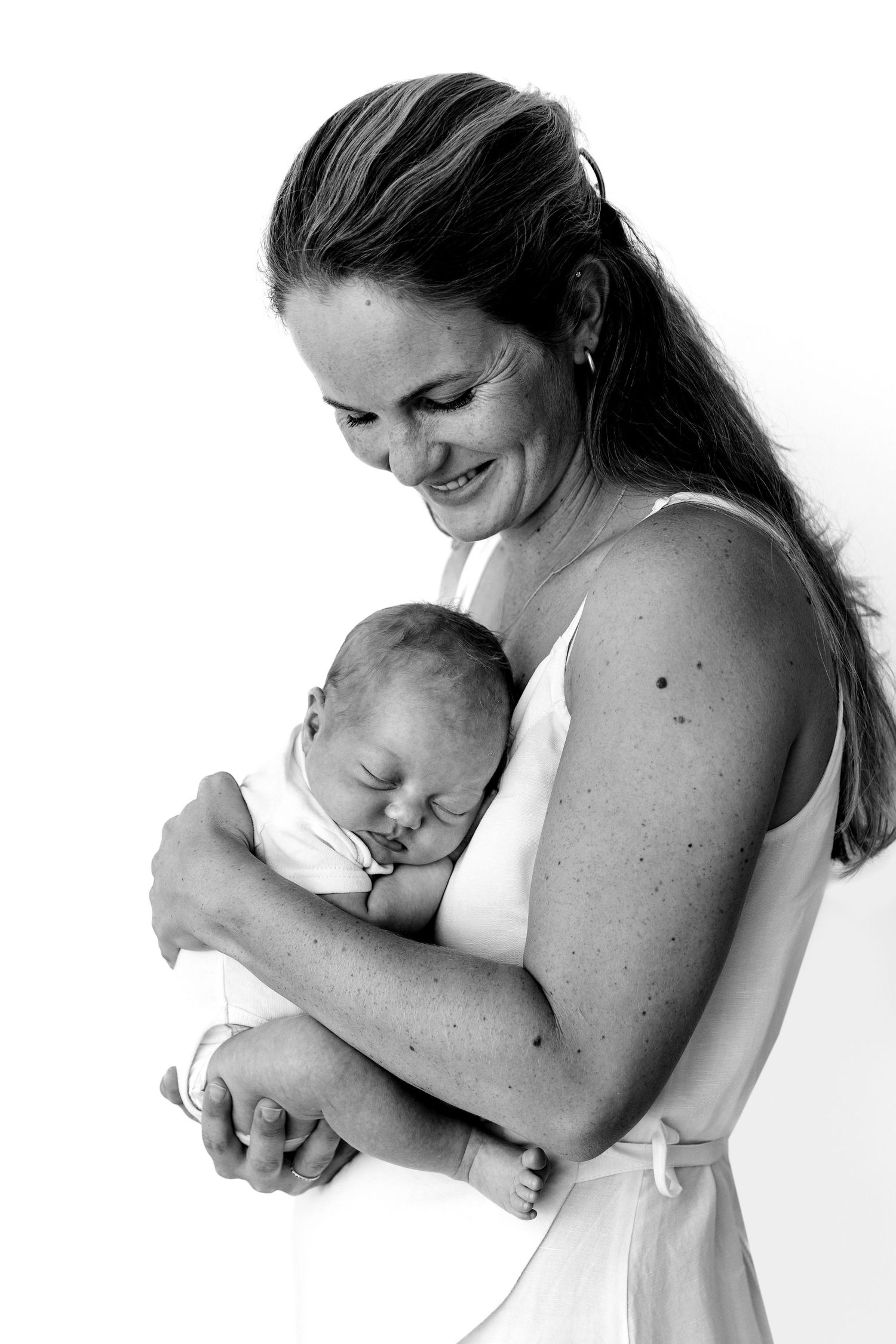 Black and white photo of a smiling mother tenderly holding her sleeping newborn baby against a white background.