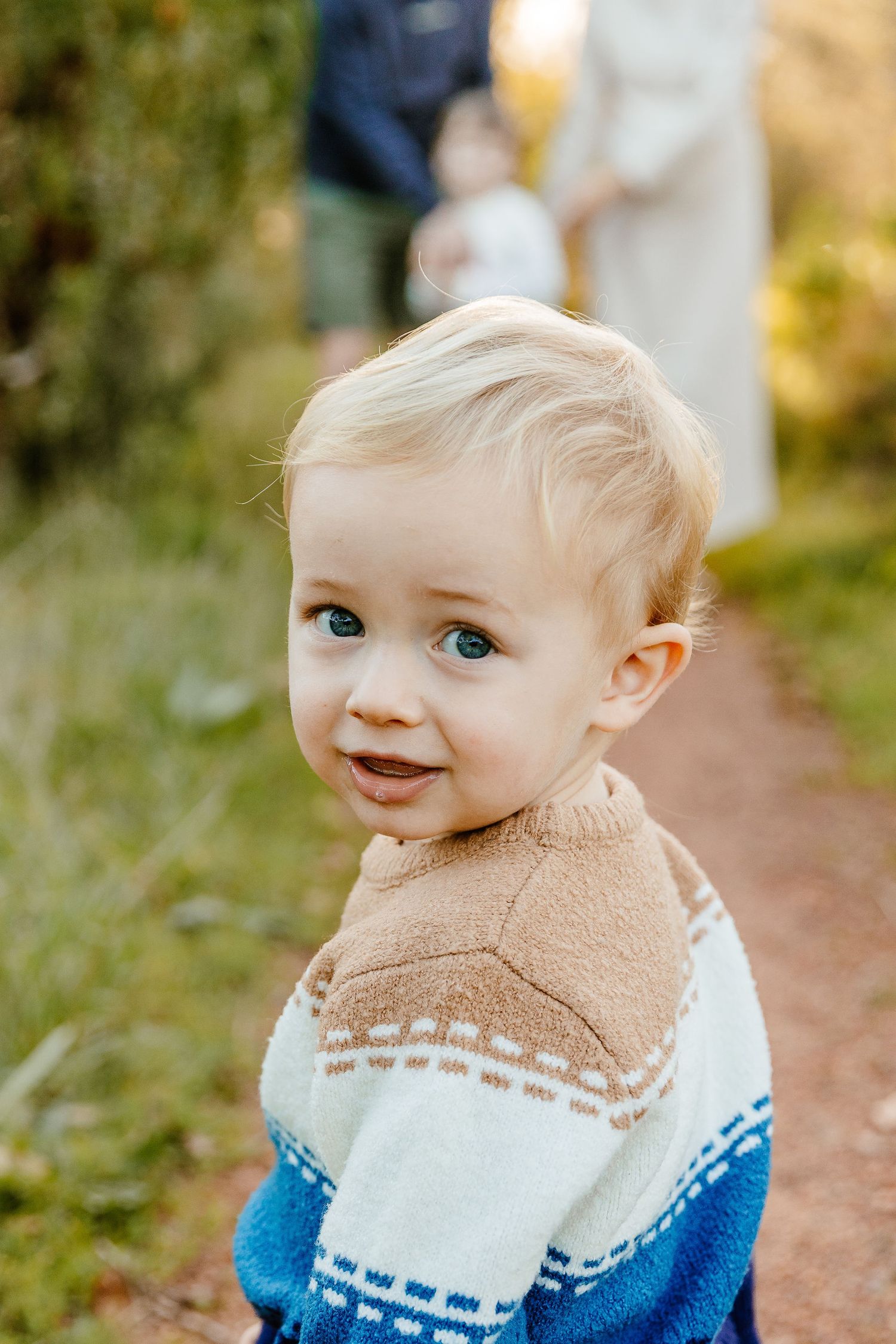 Adorable blonde toddler in patterned sweater smiling over shoulder on garden path.
