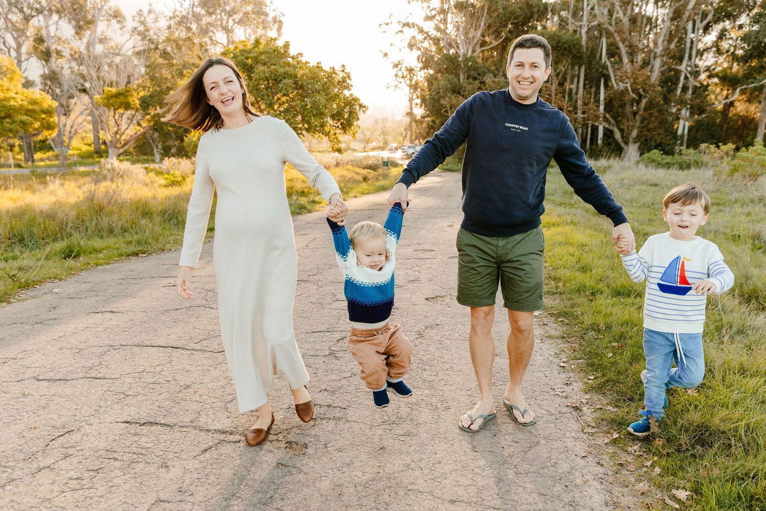 A joyful family of four holds hands walking down a sunlit dirt path surrounded by trees.
