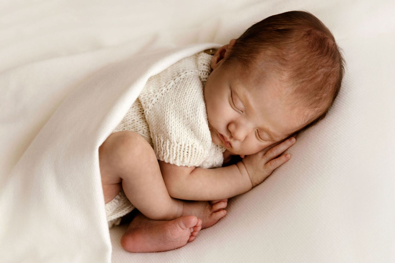 Adorable newborn baby sleeping curled up in a knit outfit tucked under a soft white blanket.