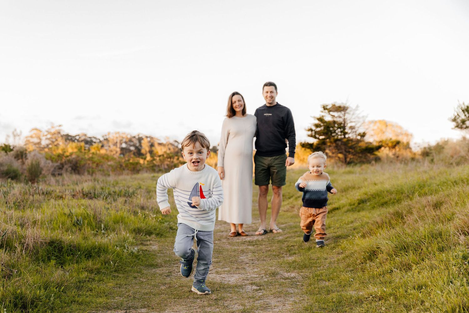 A family of four stands on a scenic meadow path as their two young boys run joyfully toward the camera.