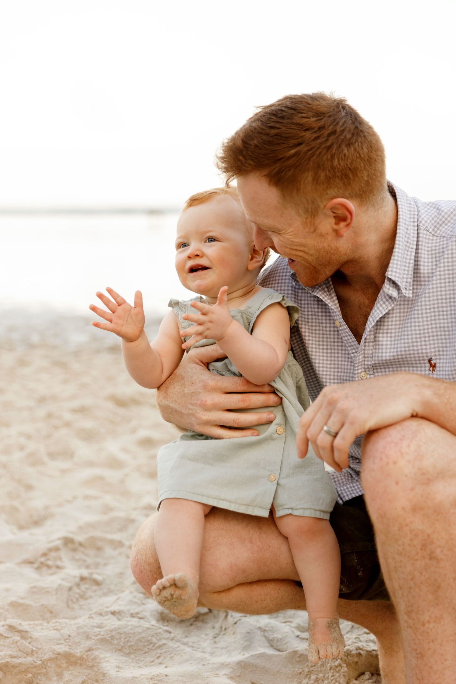 Red-haired father holding smiling baby on sandy beach, enjoying a sunny family moment by the ocean.