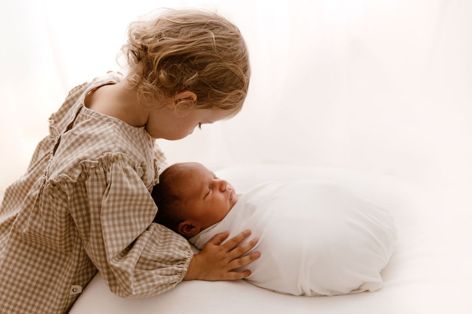 Older child in gingham dress tenderly gazing at swaddled newborn baby lying on white surface.
