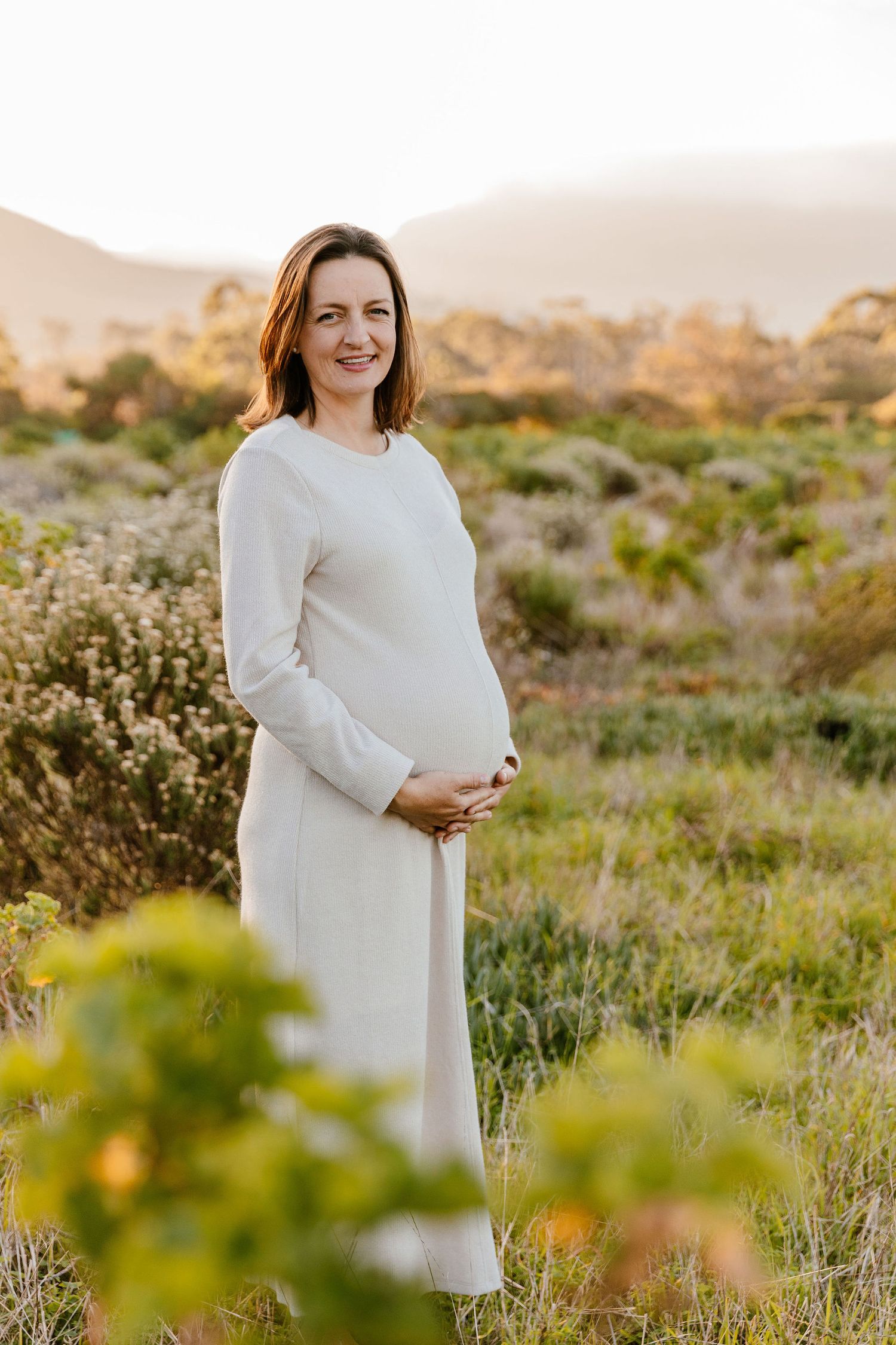 Pregnant woman in white dress cradling baby bump outdoors in golden hour light with mountains in background.