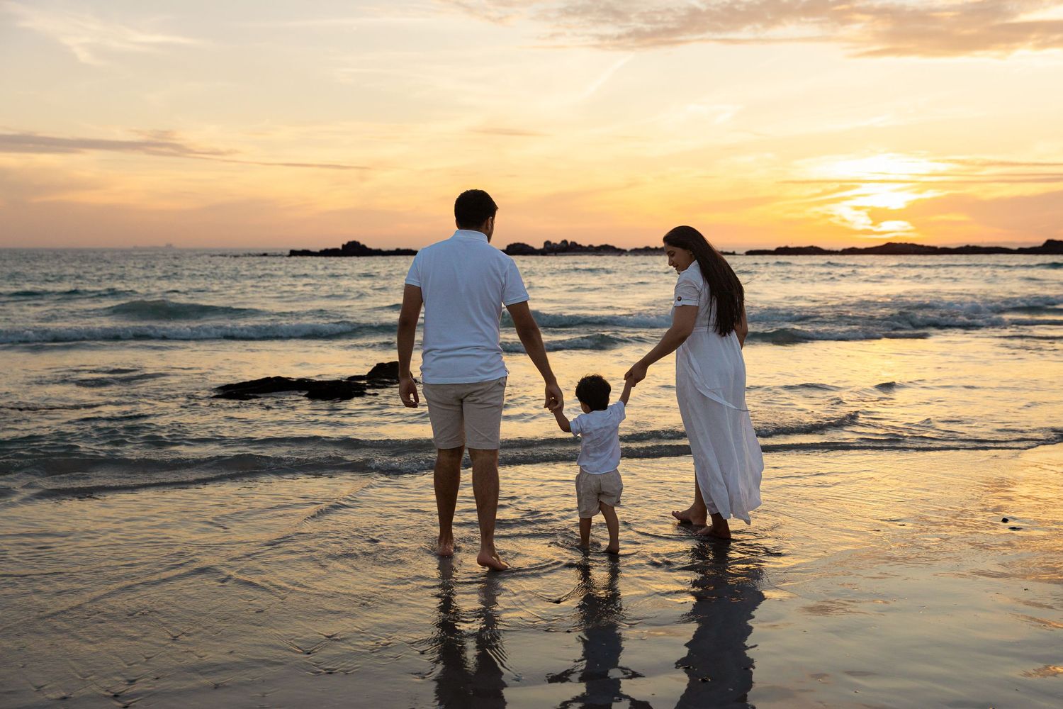 Family of three holding hands on beach at sunset, parents and toddler in white outfits enjoying ocean waves.