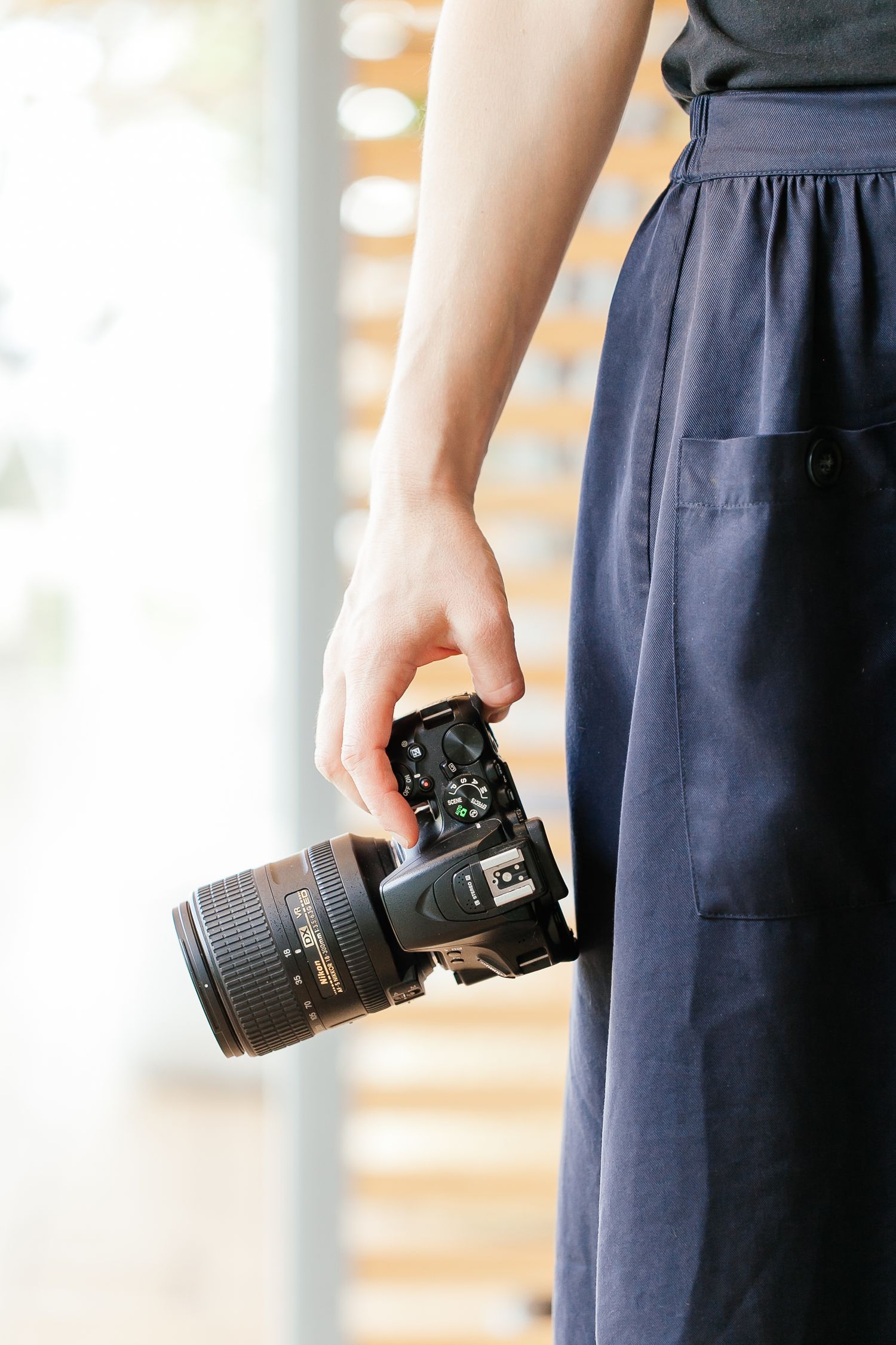 Woman in navy blue skirt holding a Nikon DSLR camera at her side indoors.