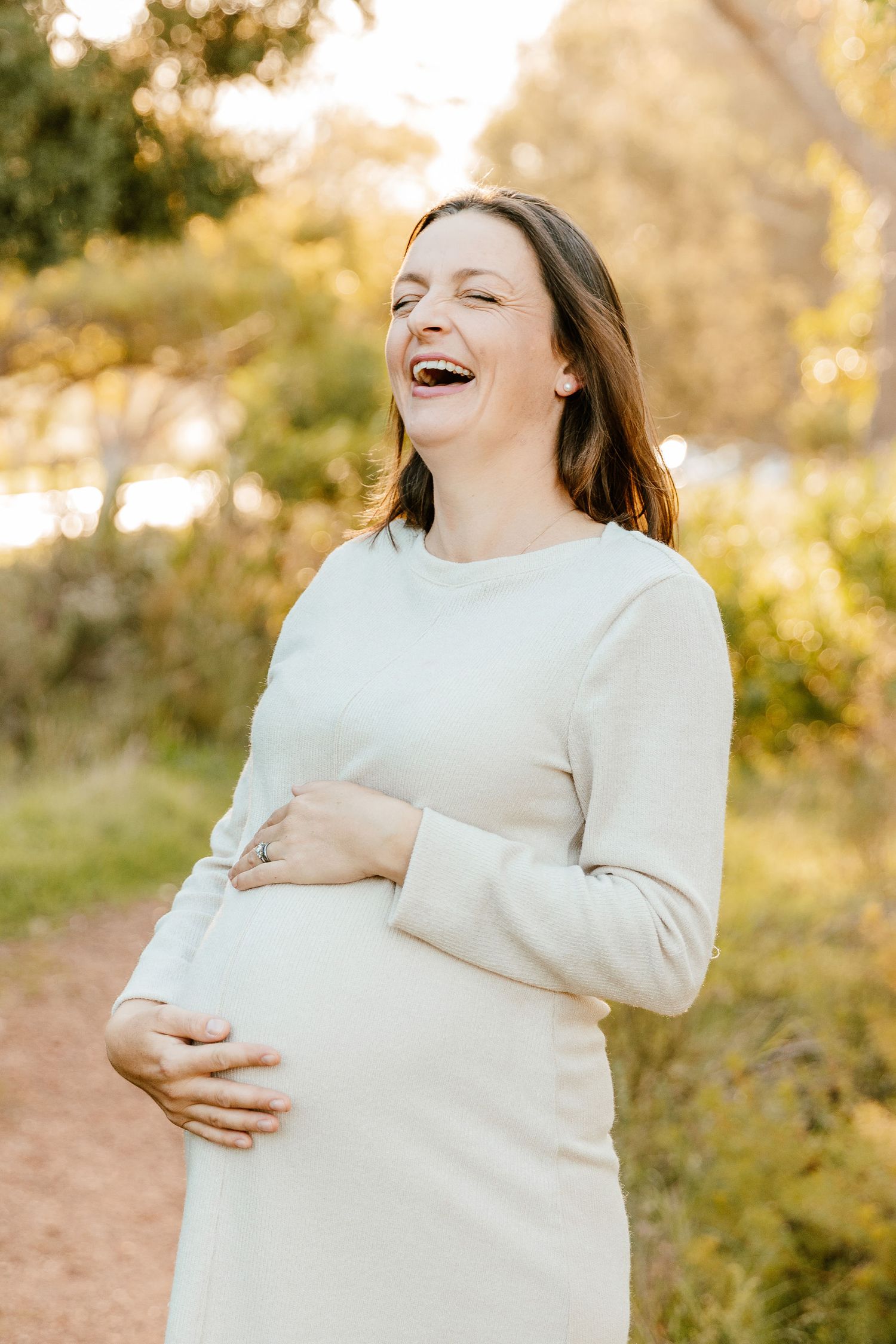 Laughing pregnant woman in white dress cradling baby bump outdoors in golden sunlight.