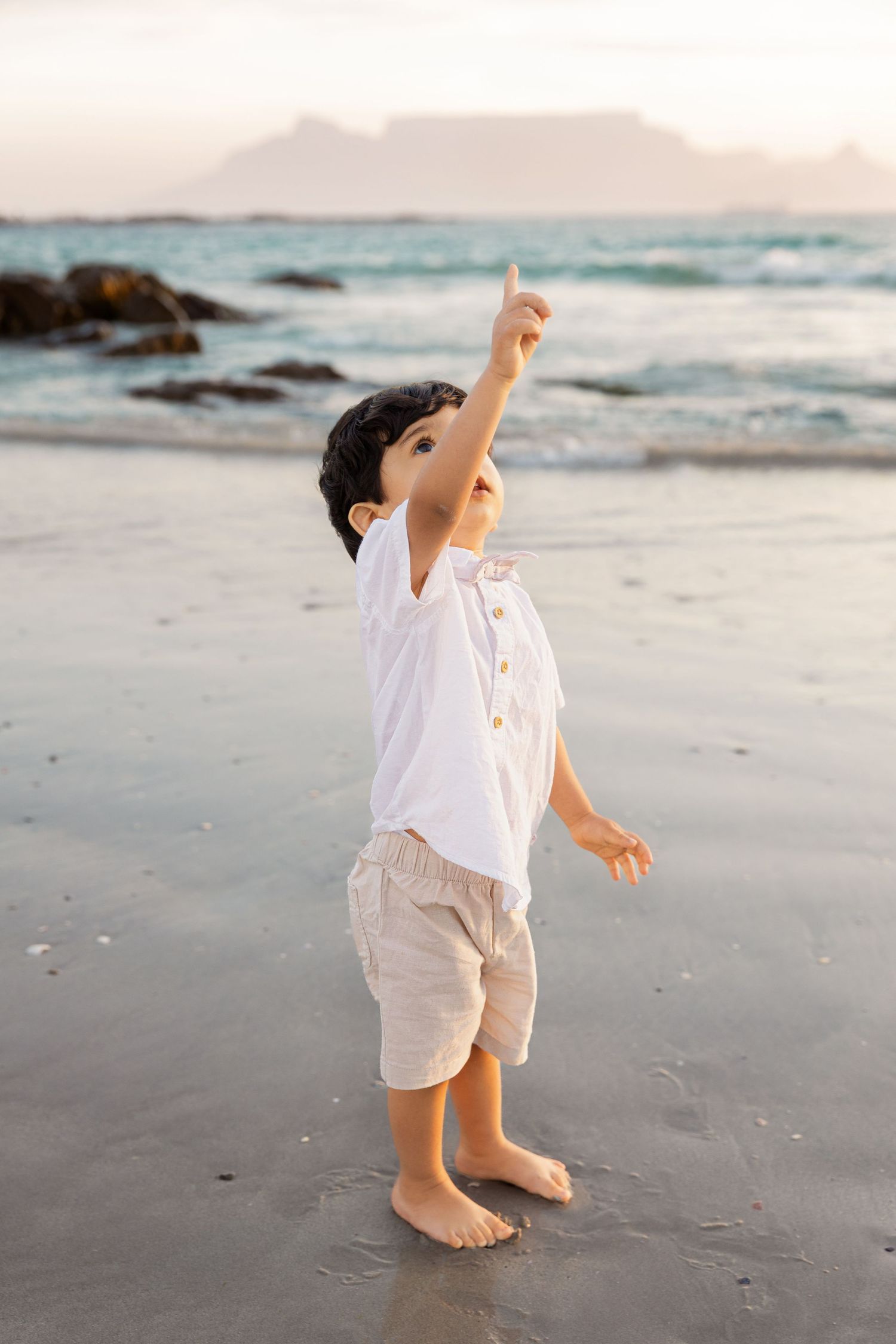 Young boy in white shirt pointing up at the sky on a sandy beach with Table Mountain in the background.