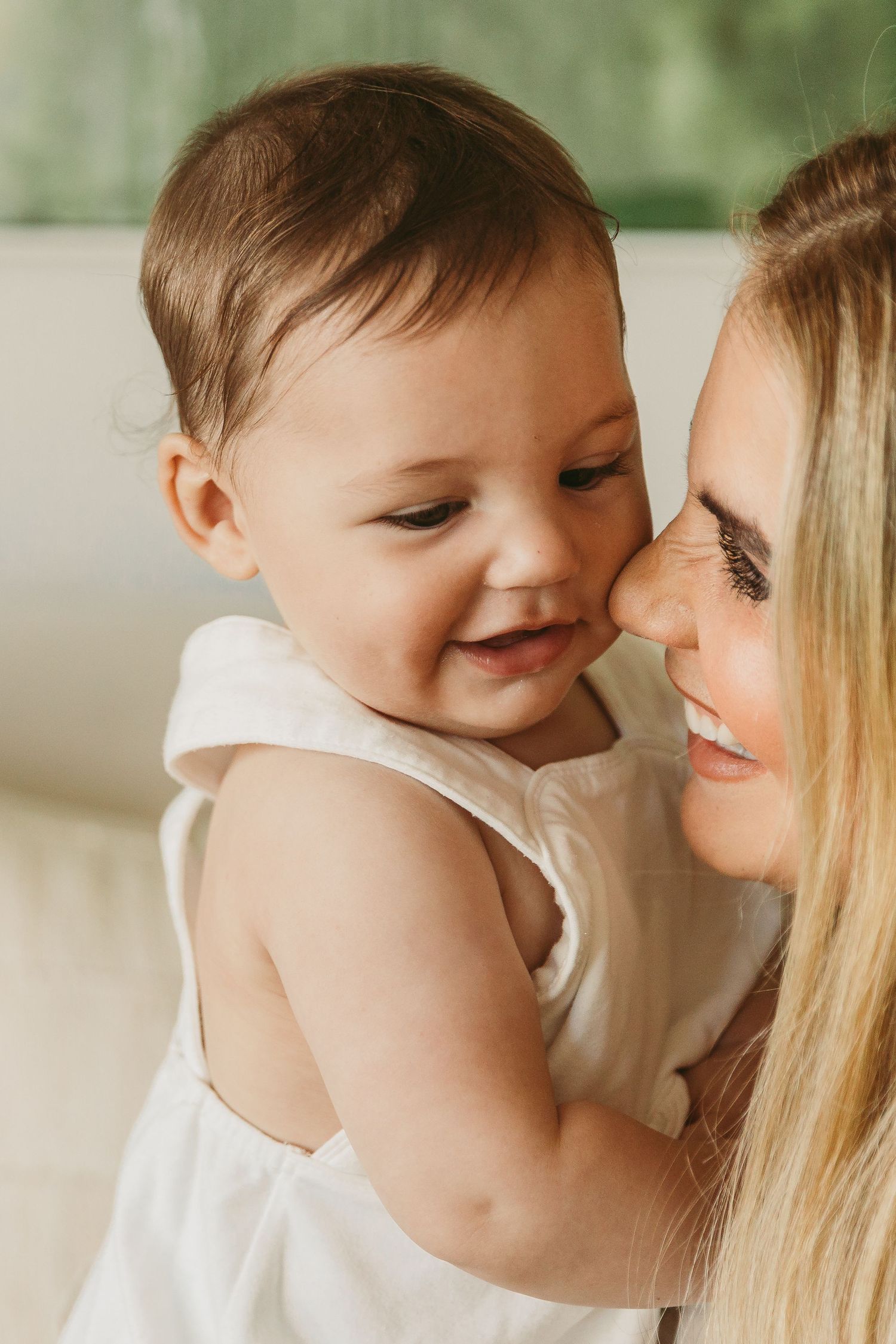 A smiling blonde mother holds her happy baby girl in a white dress, sharing a tender nose-to-nose moment.