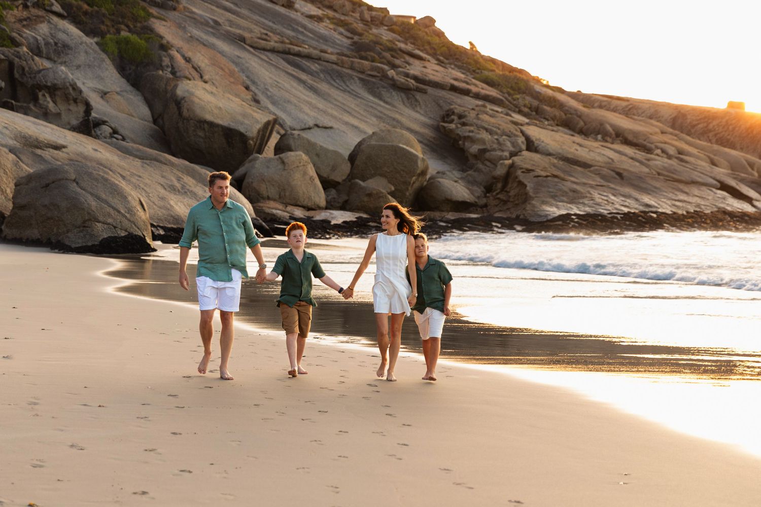 Family of four holding hands walking on a sandy beach at sunset with rocky cliffs in the background.