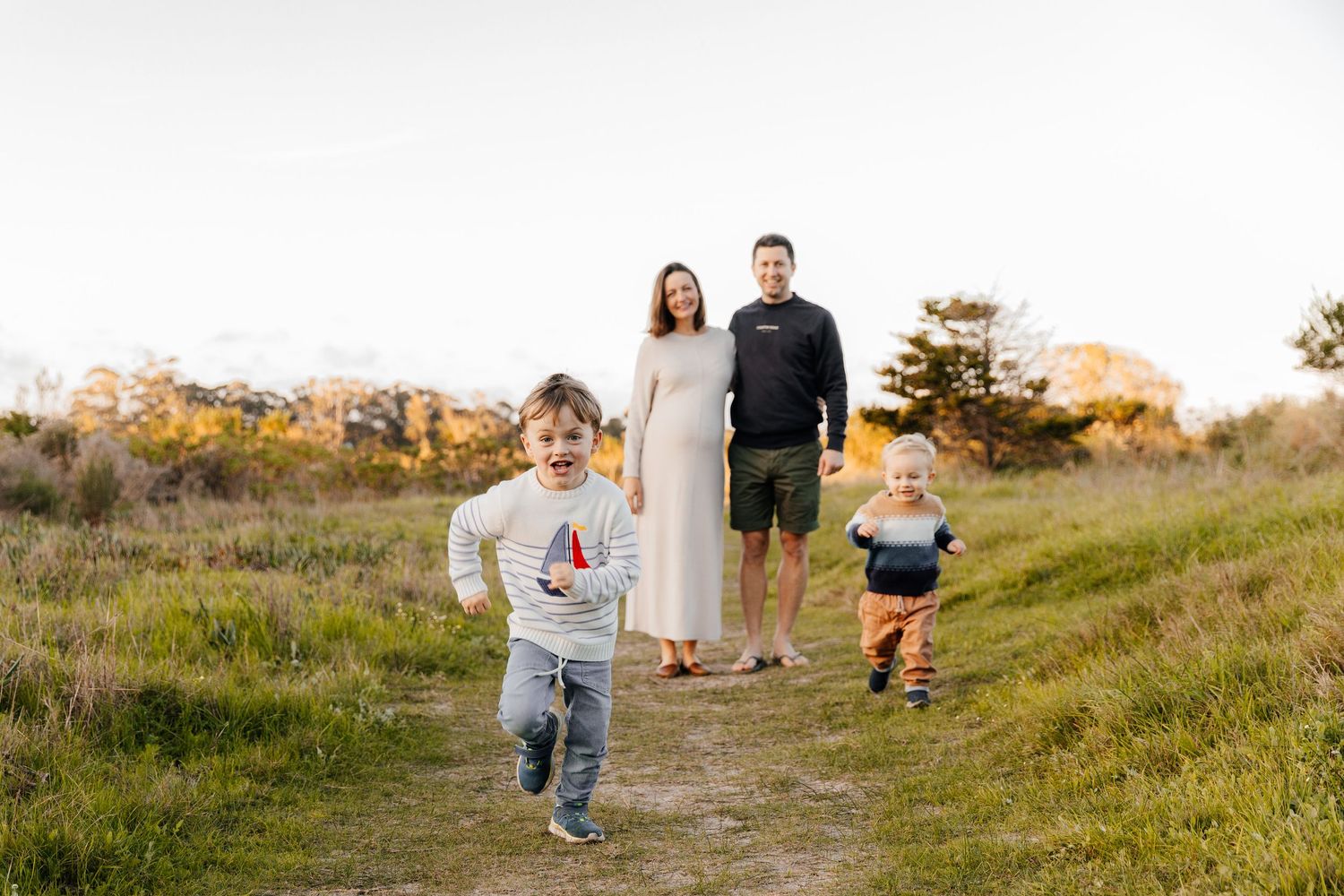 Family of four enjoying a sunny outdoor walk, two young boys running ahead of their smiling parents on a grassy path.
