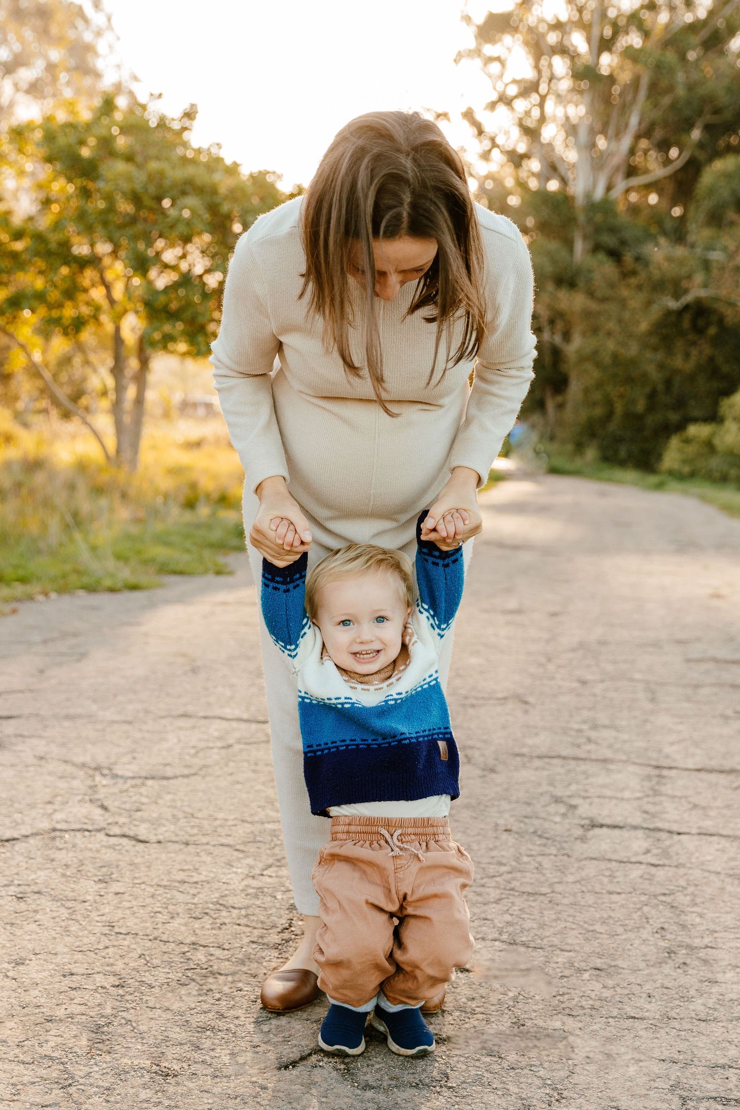 A pregnant mother gently holds her smiling toddler son's hands on a warm sunlit dirt path.