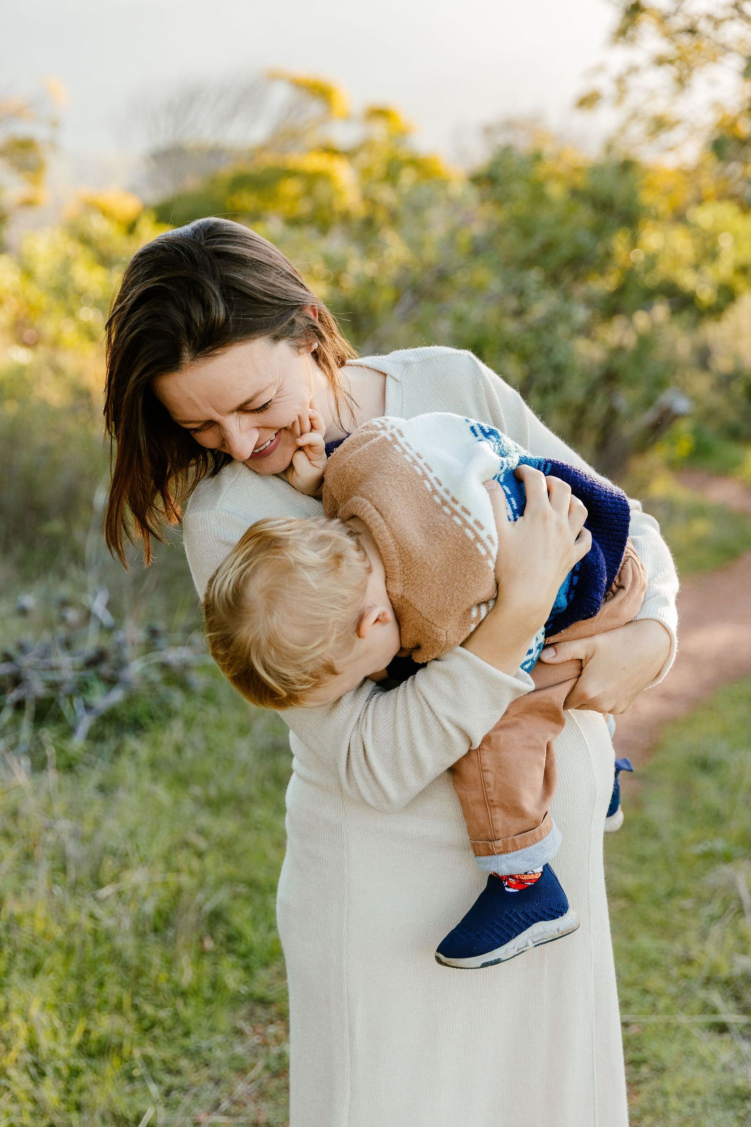 Mother in white dress laughing joyfully while holding her young toddler son outdoors.