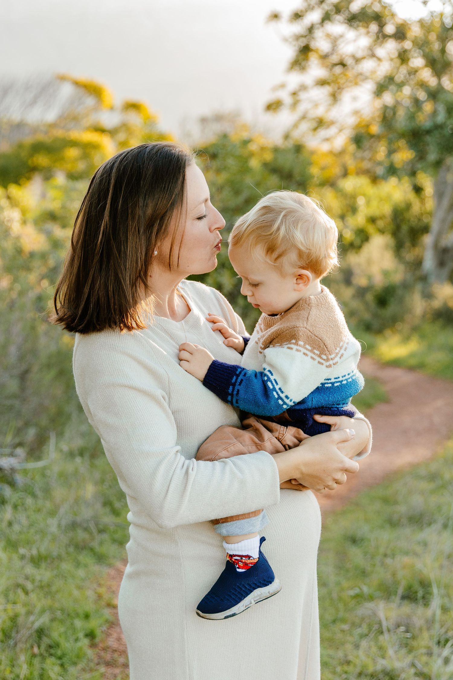 Pregnant mother in white dress tenderly kissing her young blond toddler outdoors.