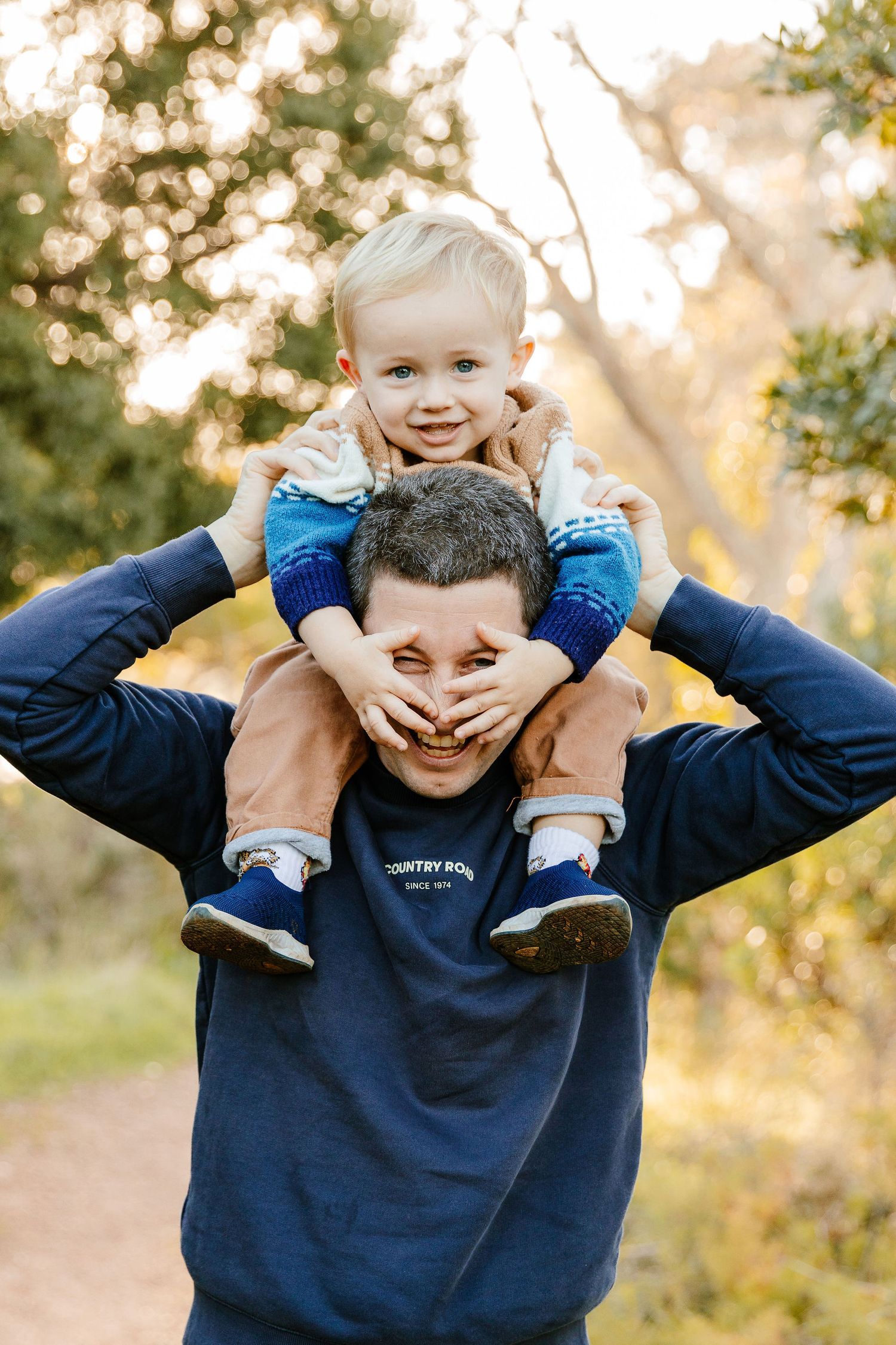 Happy father carrying young blonde toddler on his shoulders in a sunlit outdoor park setting.