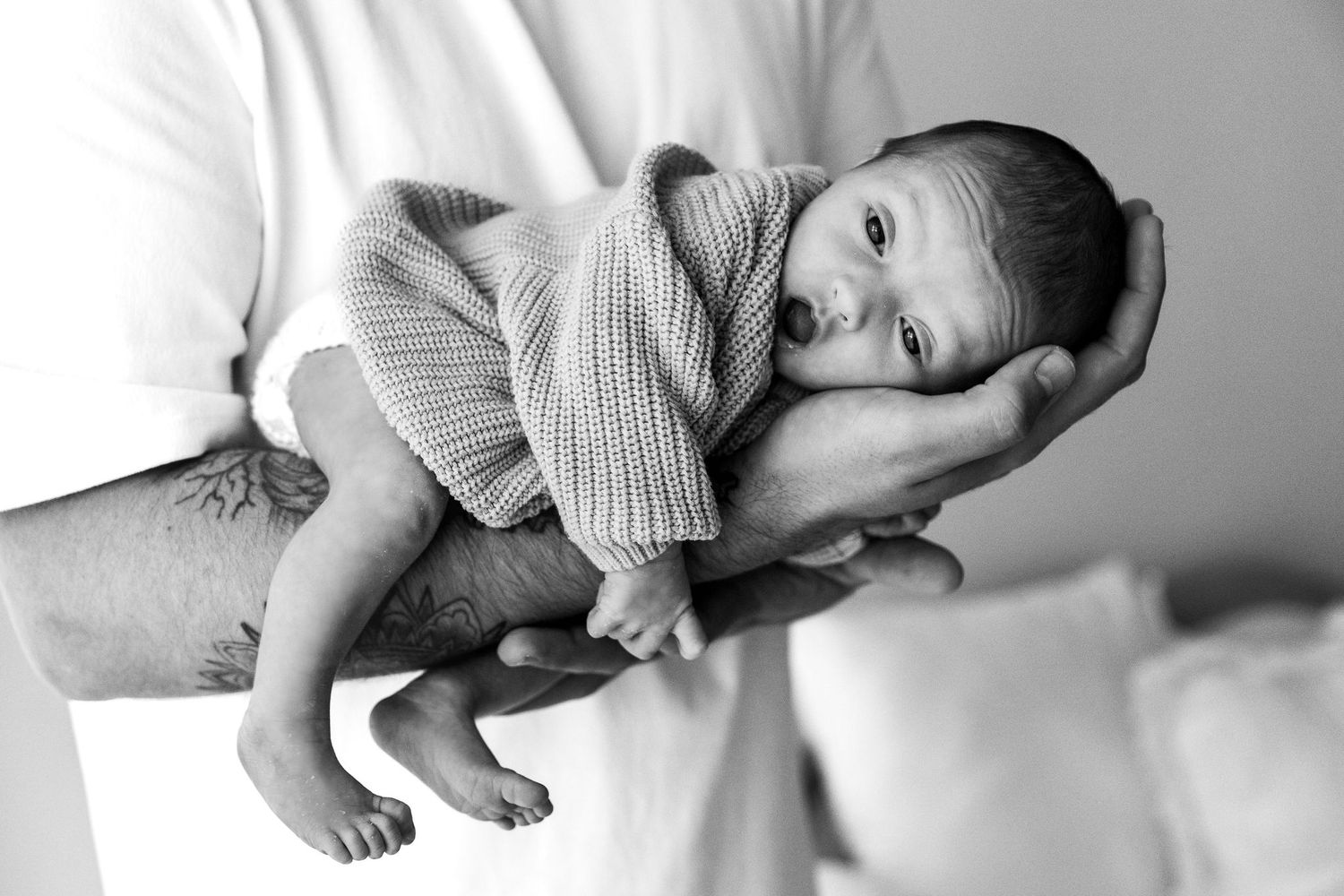 Black and white photo of father with tattoos holding alert newborn baby in a brown knit outfit.