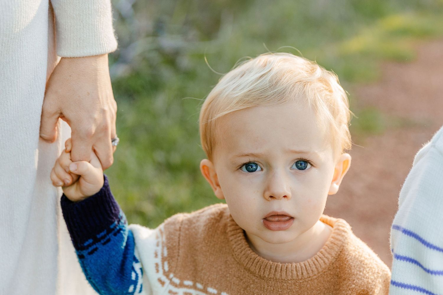 Close-up portrait of a blue-eyed blonde toddler holding a parent's hand outdoors.