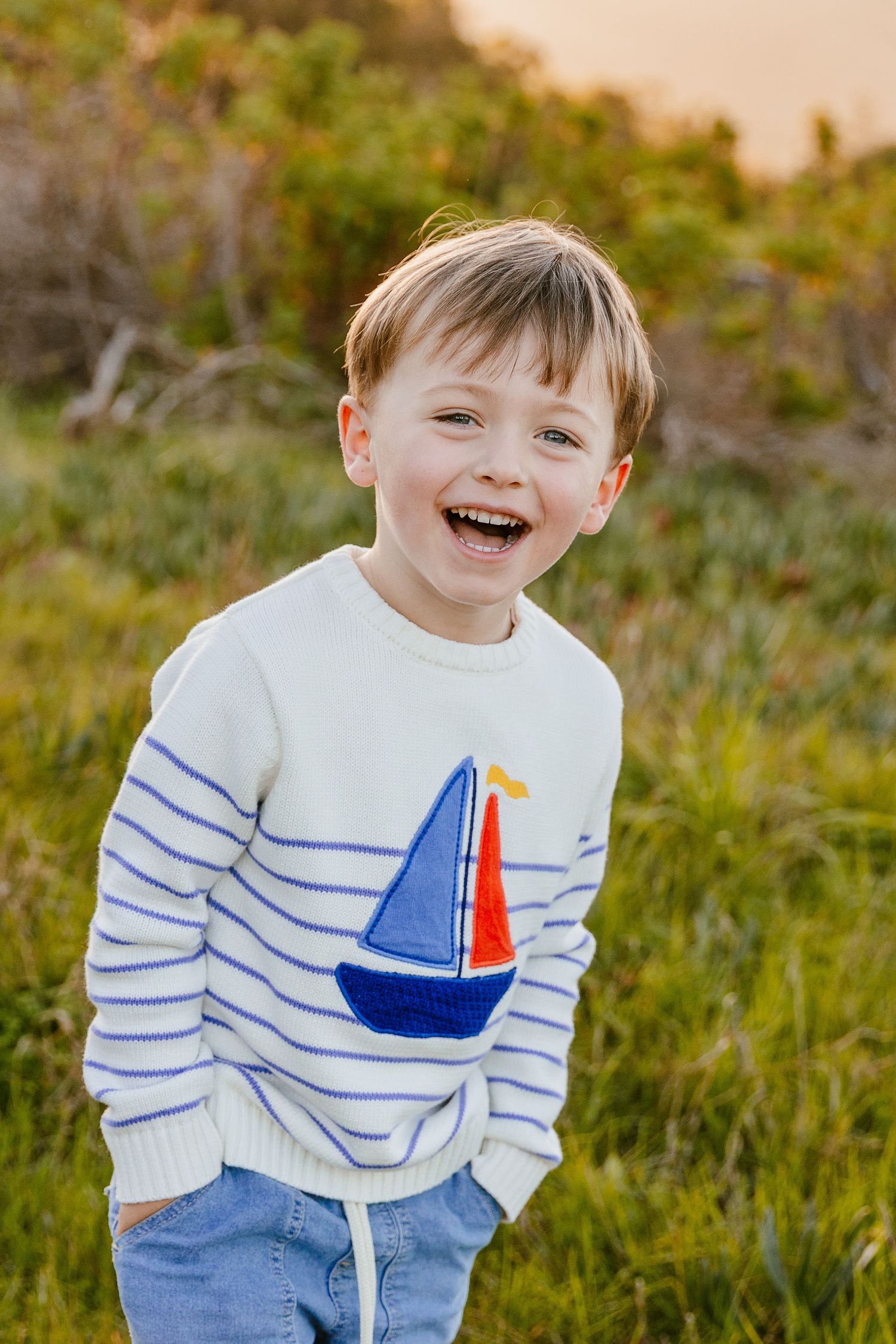 Laughing young boy in a striped sailboat sweater standing in a lush green field.