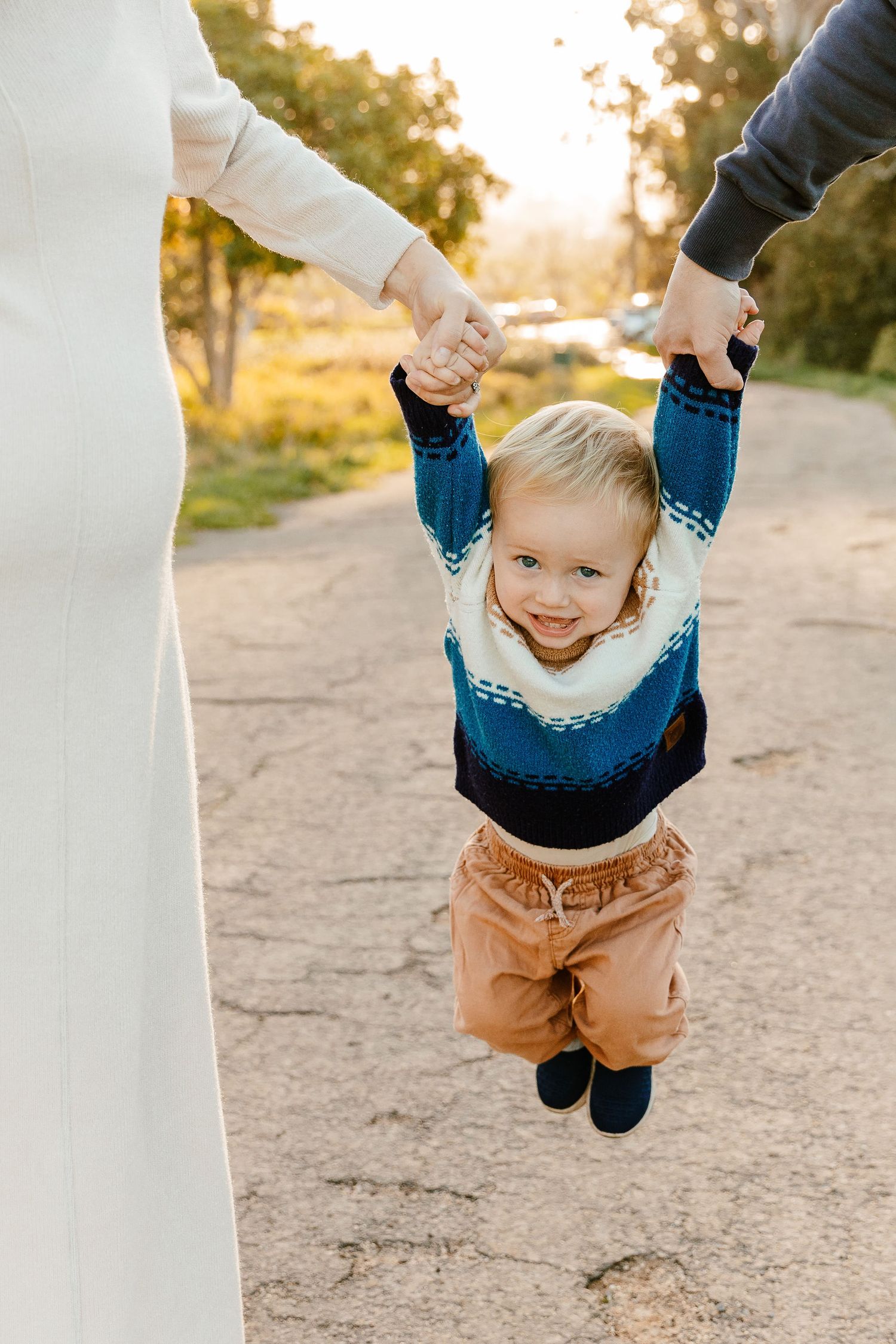 Joyful toddler boy being swung by parents hands on a sunny outdoor path during a family photo session.