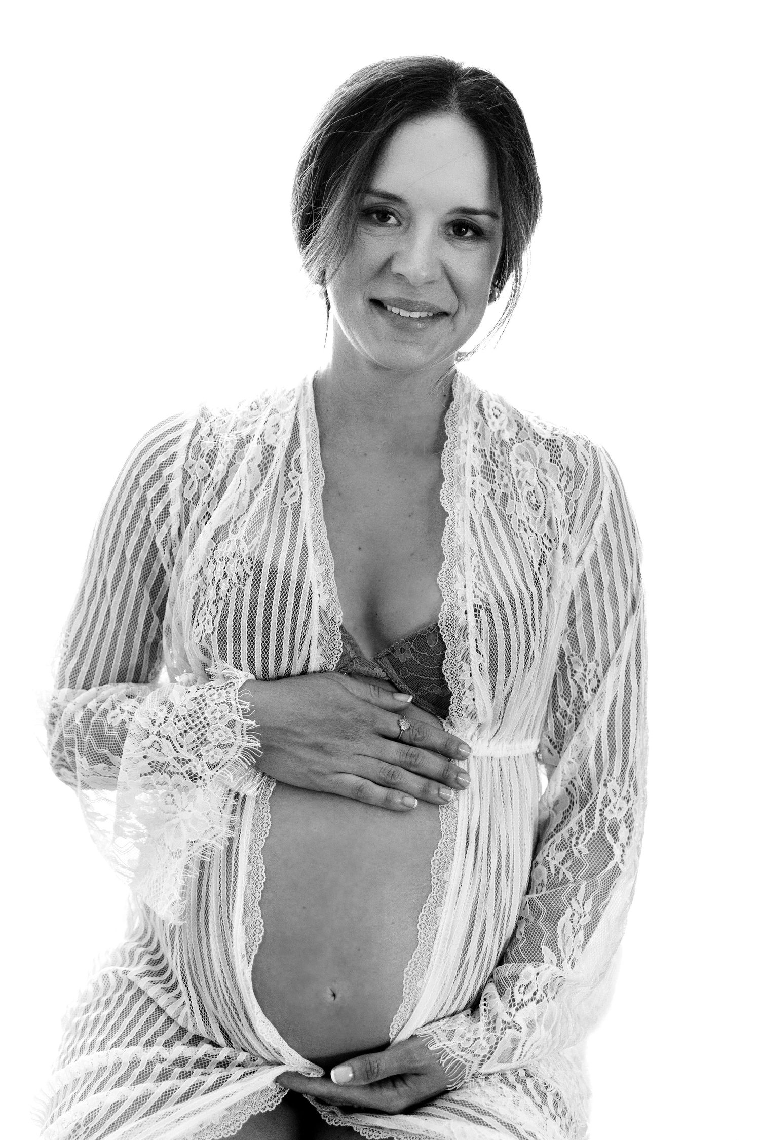 Black and white portrait of a smiling pregnant woman in lace robe cradling her baby bump.