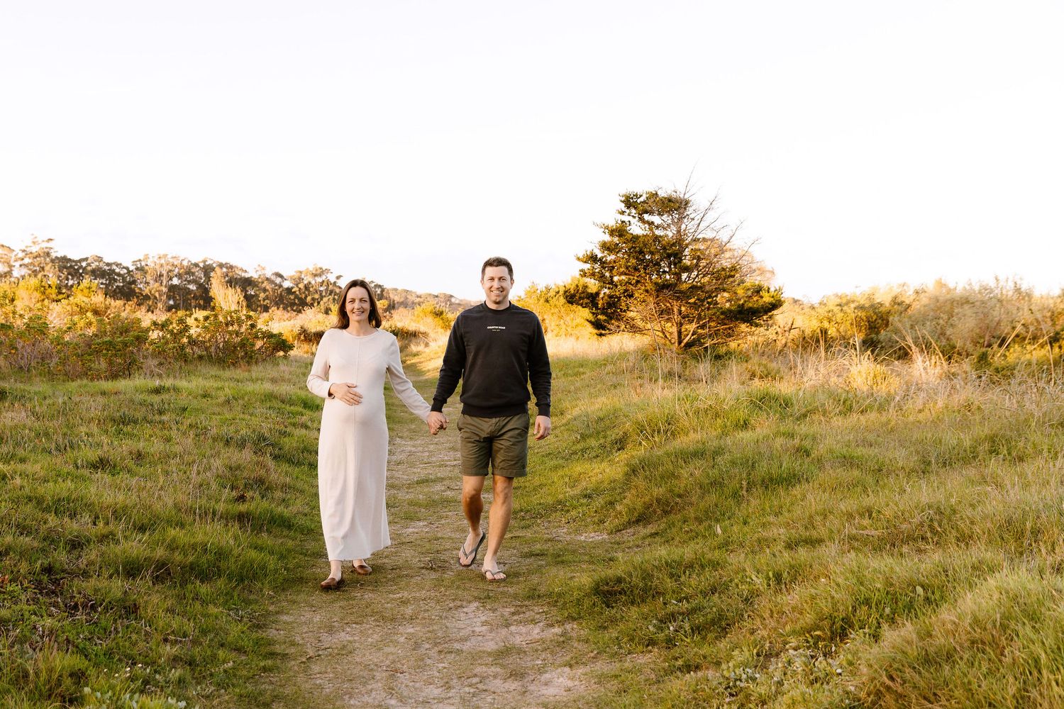 Couple holding hands walking on a sunlit path through golden meadow, woman in white dress with baby bump.