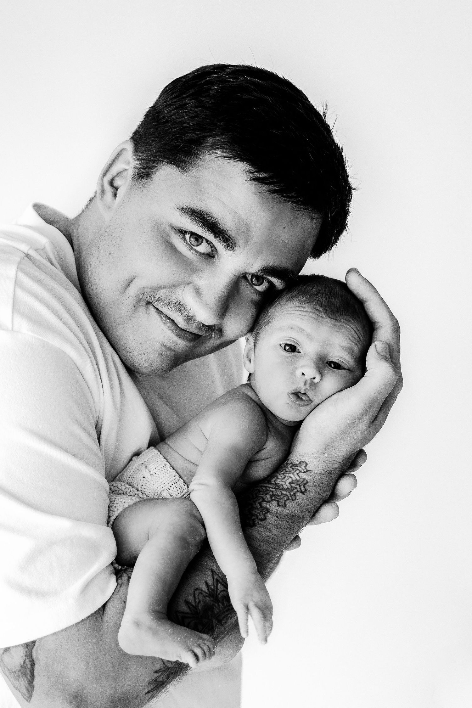 Black and white portrait of smiling tattooed father holding his alert newborn baby.