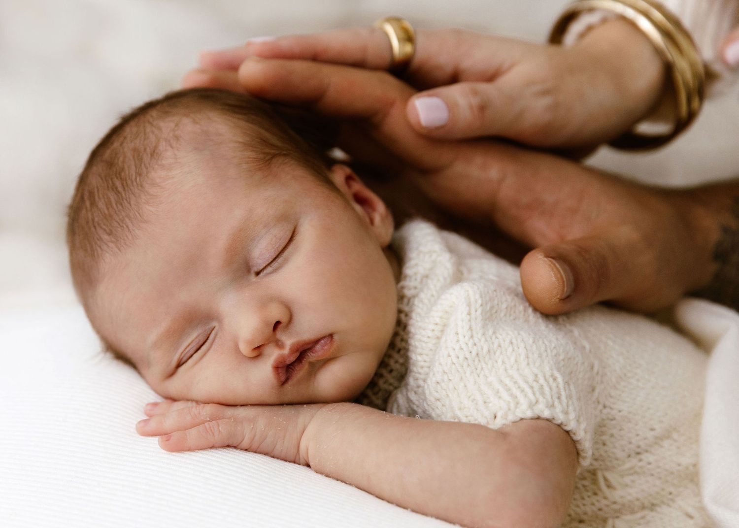 Close-up of peaceful sleeping newborn baby in white knit outfit with parent's hand gently touching head.