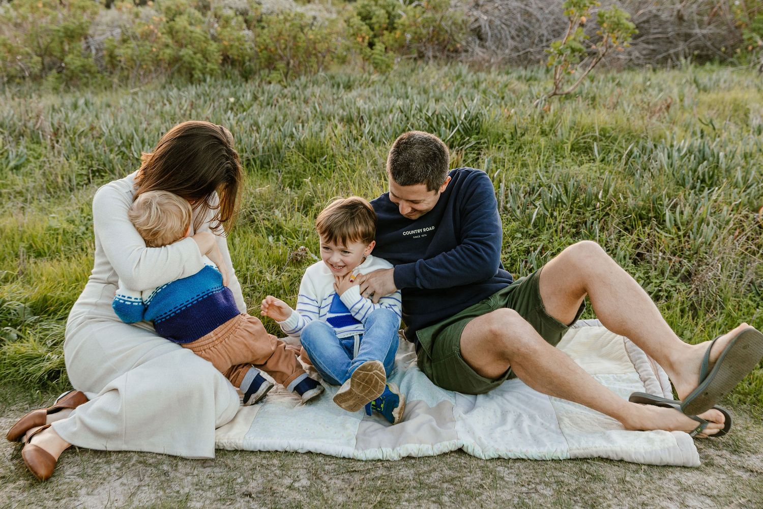 Family of four sitting closely together on a blanket in a grassy outdoor setting.