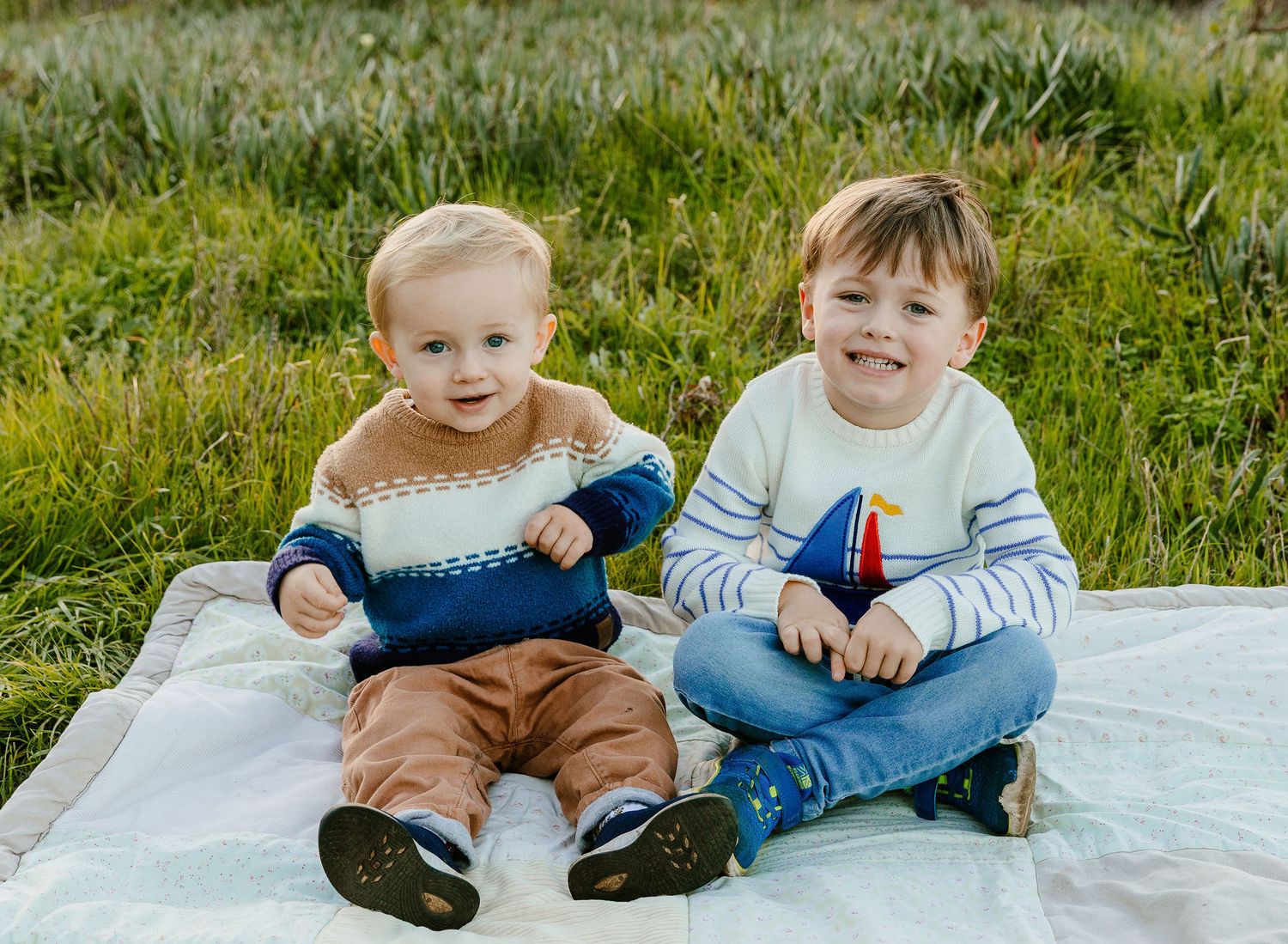 Two young brothers sitting together on a blanket in a grassy field smiling at camera.