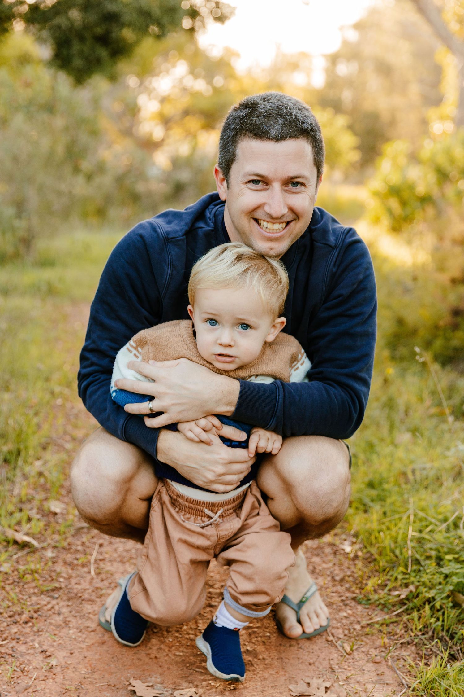 Smiling father crouching outdoors hugging his blonde toddler son in a sunny nature setting.