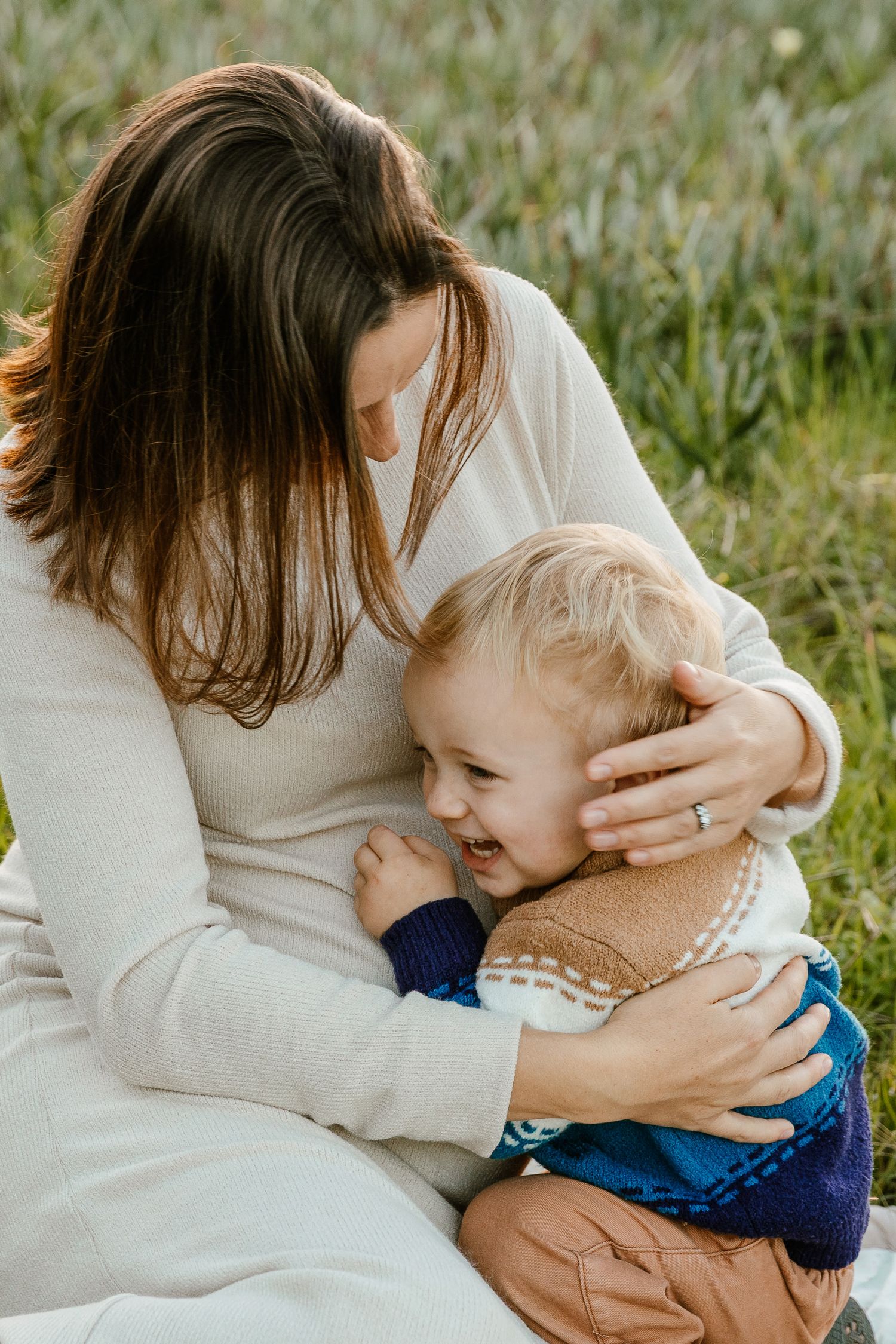 A mother tenderly embraces her laughing toddler while sitting outdoors in a lush green field.