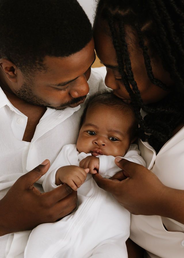 Two parents lovingly kiss their newborn baby dressed in white during a tender family portrait session.