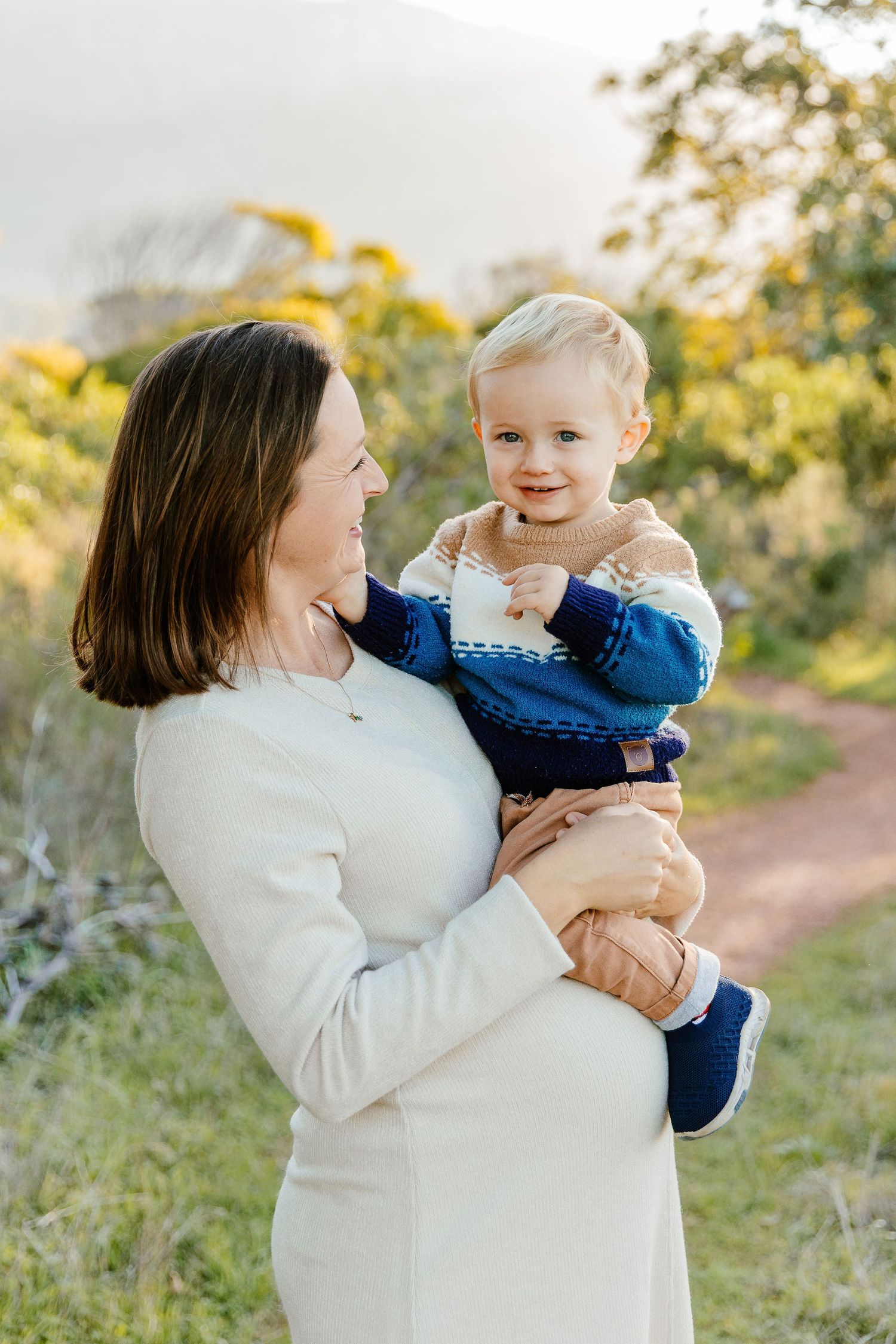 Pregnant woman in white dress lovingly holding a smiling blonde toddler outdoors.