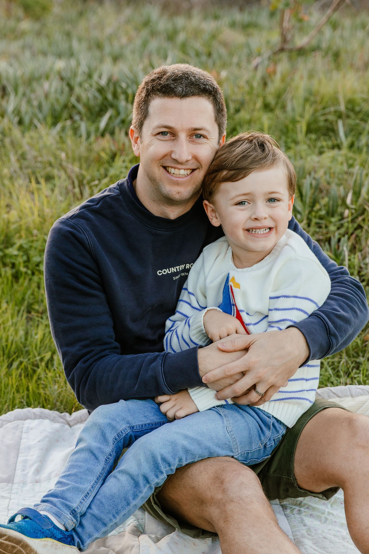 A smiling father hugs his happy young son while sitting on a blanket in a green grassy field.