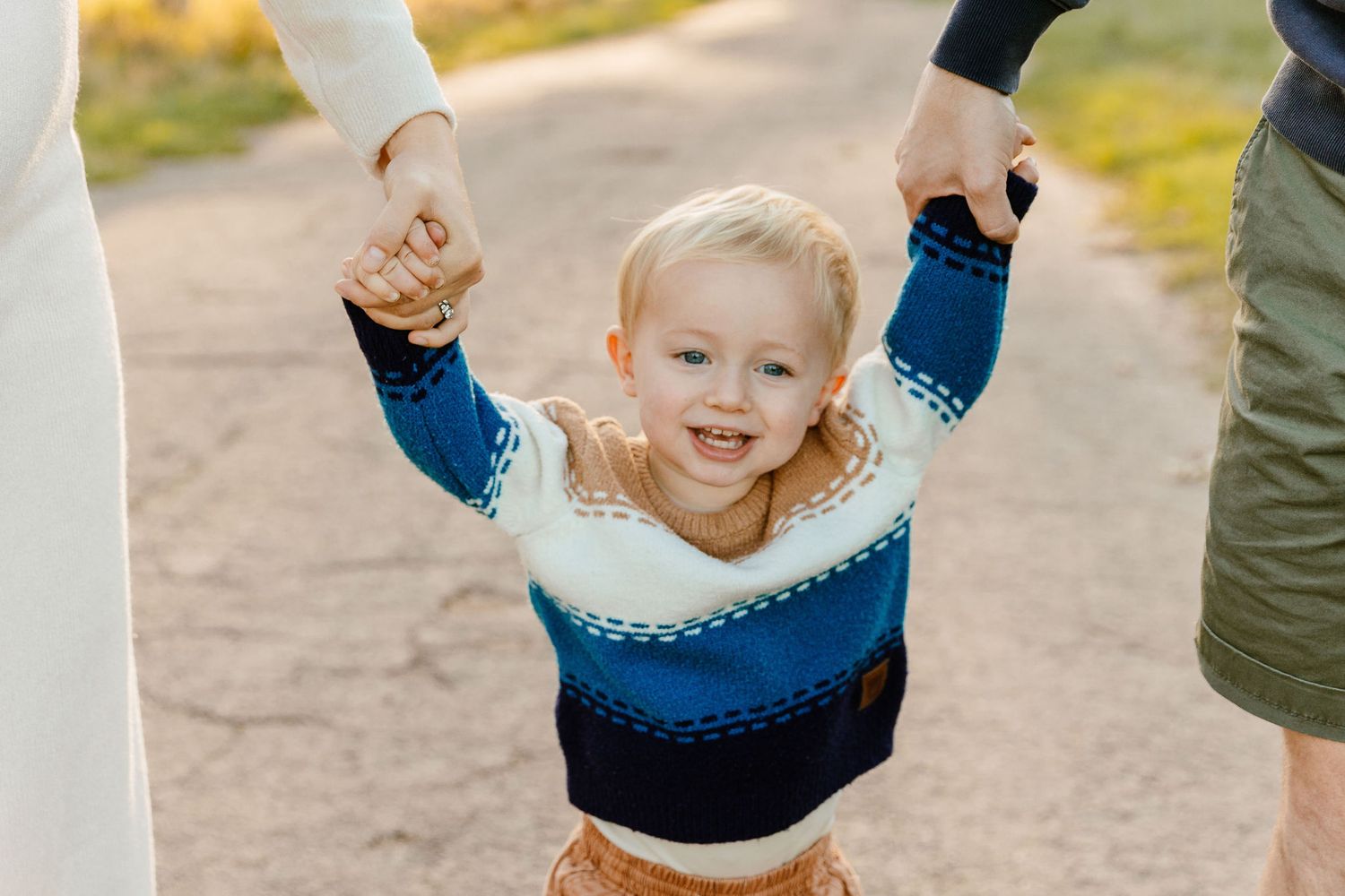 Cheerful toddler in blue sweater held by both hands by parents outdoors.