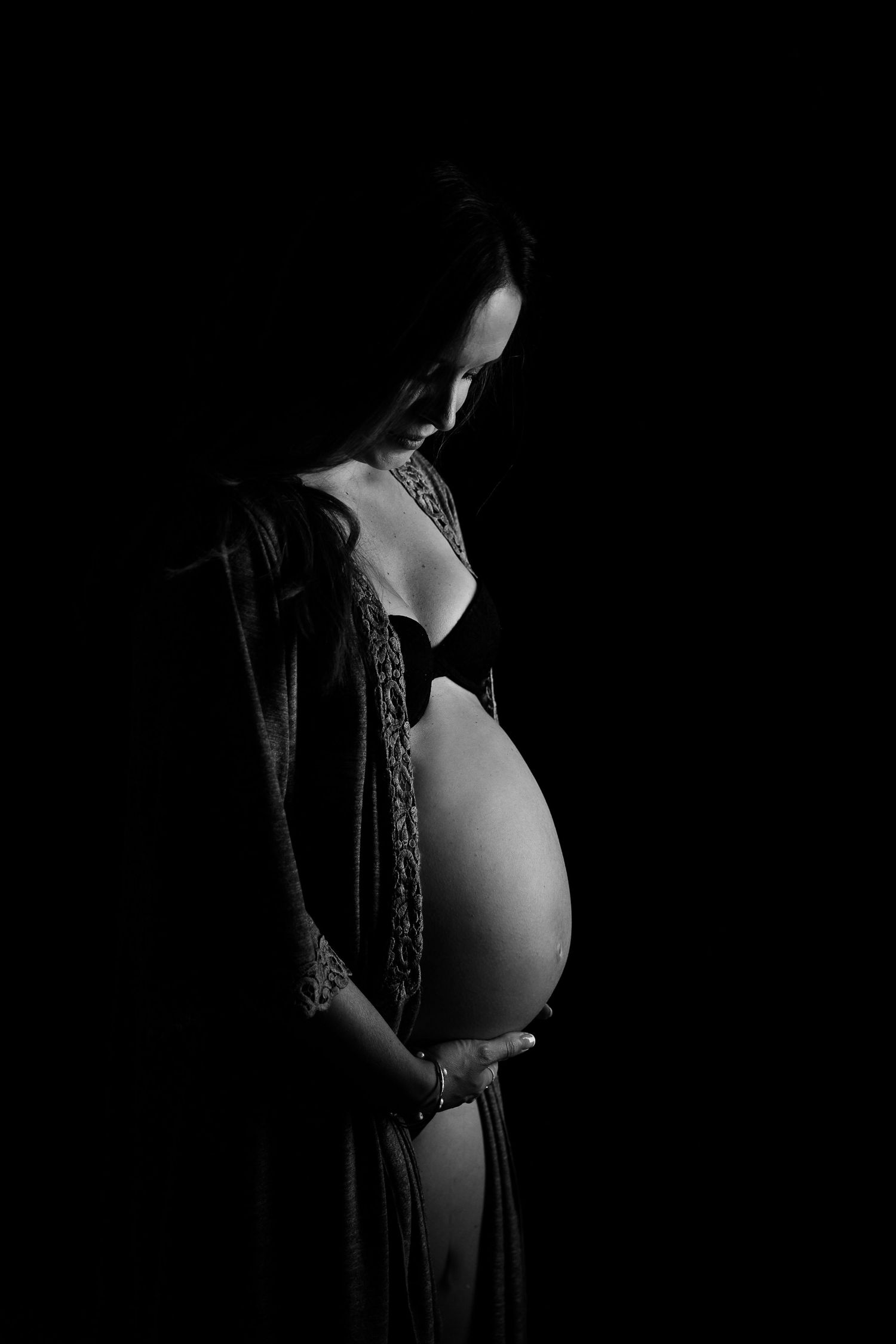 Black and white maternity photo of pregnant woman cradling baby bump against dark background.
