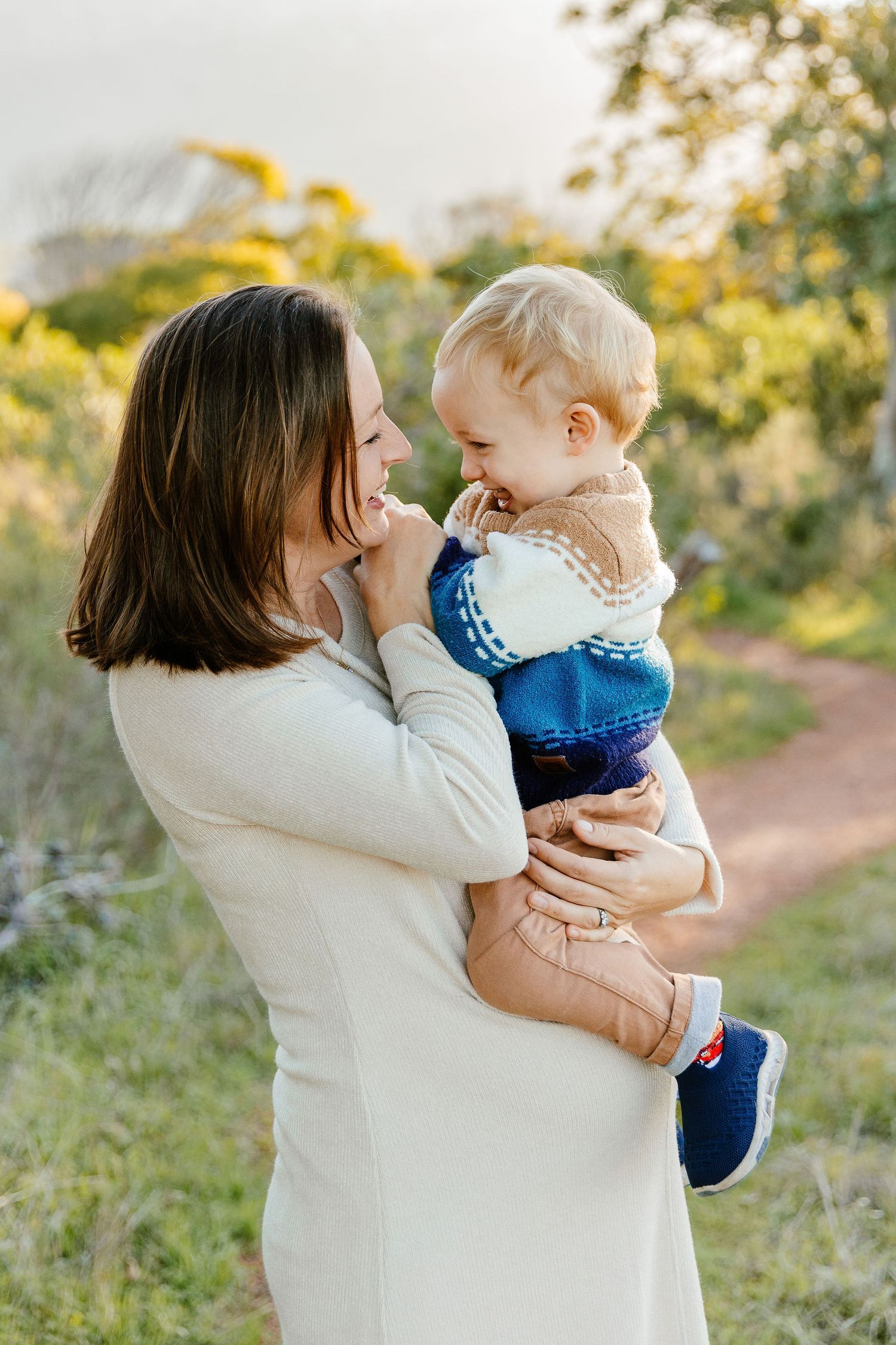 A smiling mother in a white dress holds her blonde toddler outdoors on a sunlit nature path.
