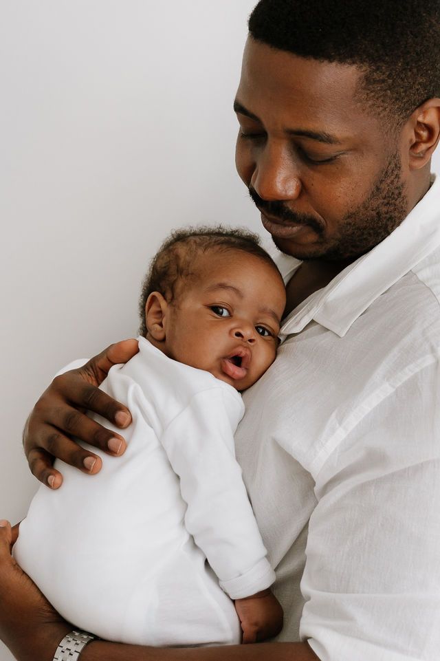 A loving father in a white shirt gently cradles his newborn baby dressed in white against a light background.