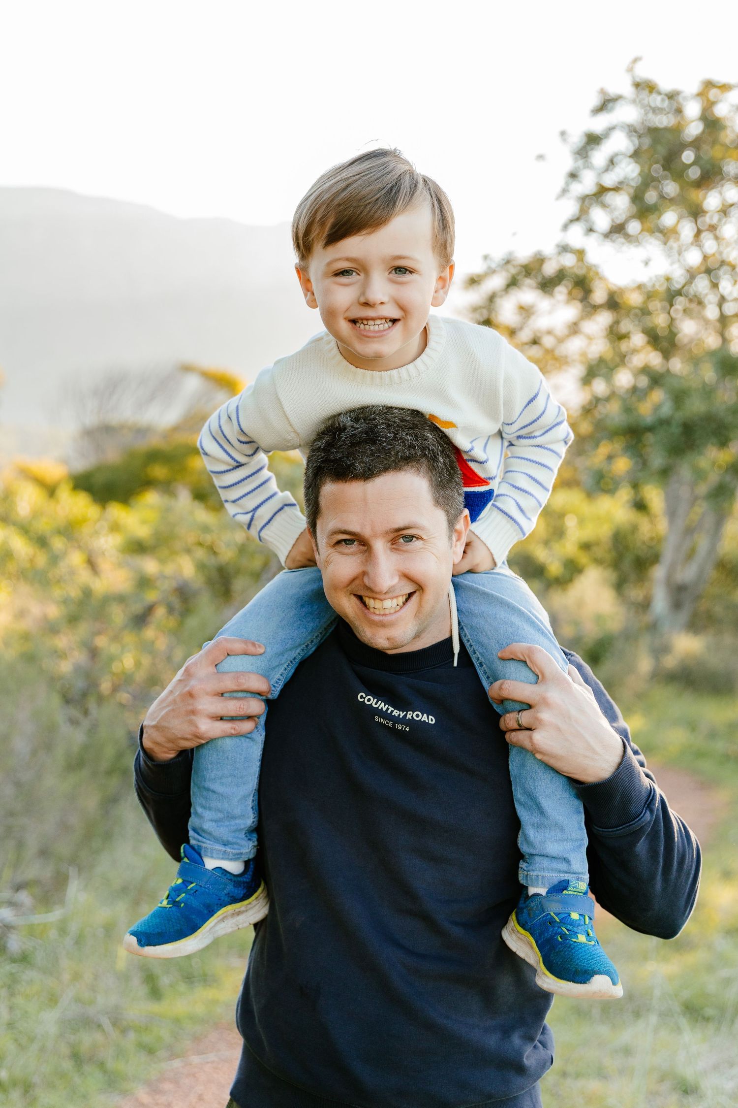 Smiling man carrying happy young boy on his shoulders outdoors on a sunny trail surrounded by greenery.