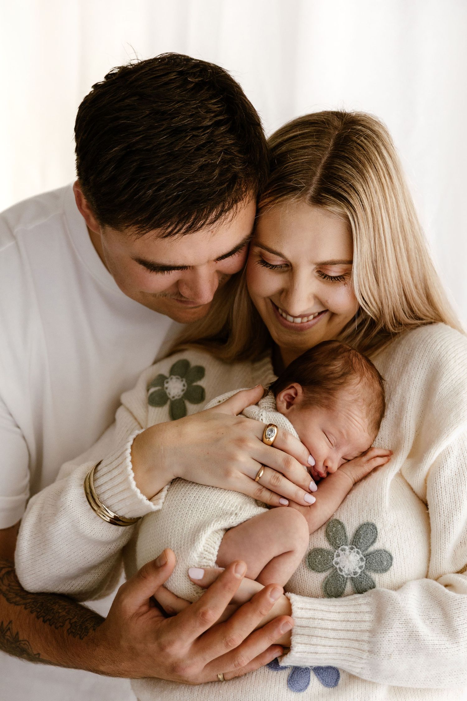 Smiling couple in white floral sweaters gently cradling their peaceful sleeping newborn baby.
