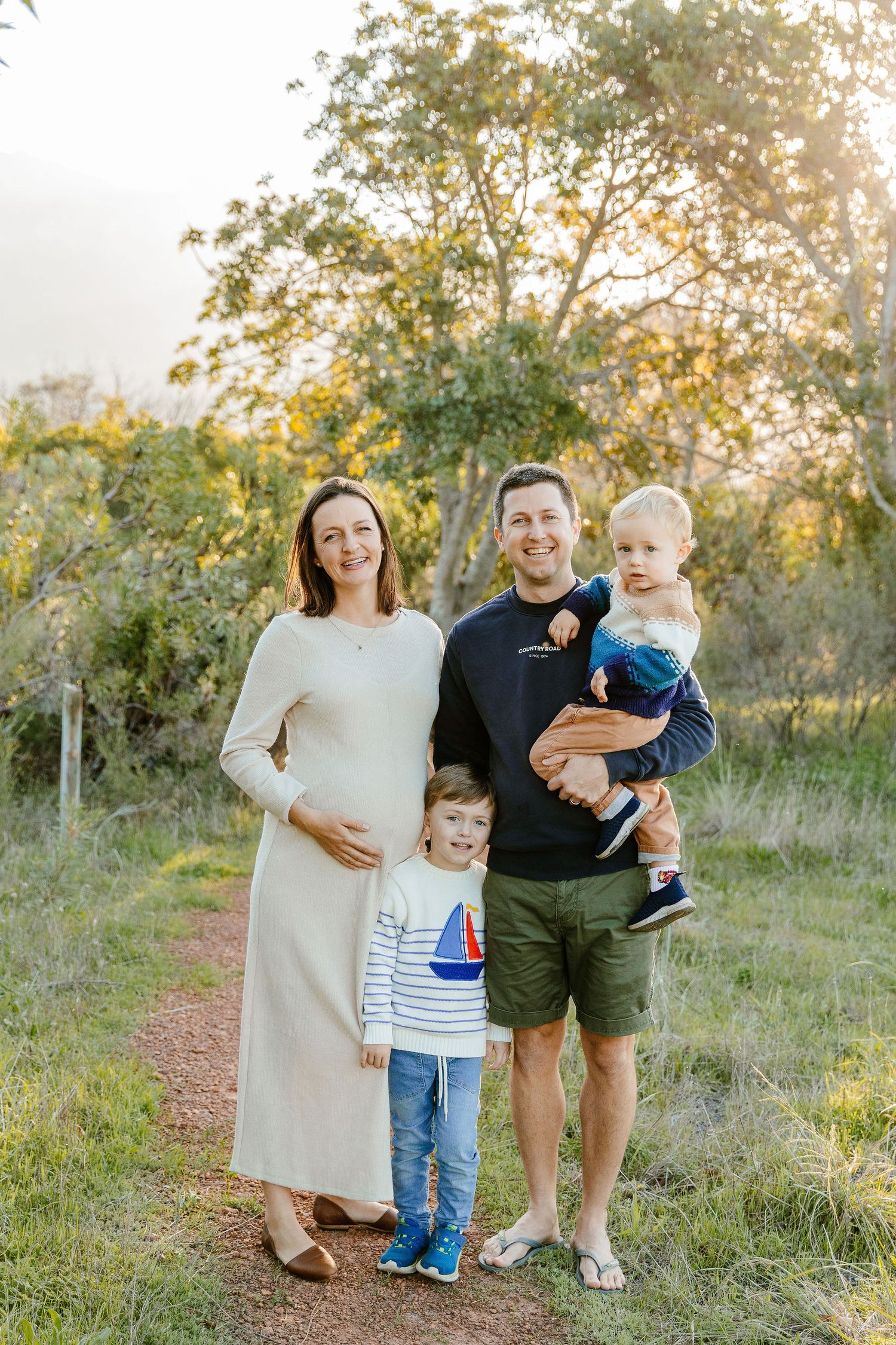 A happy pregnant family of four poses on a nature trail during golden hour sunset.