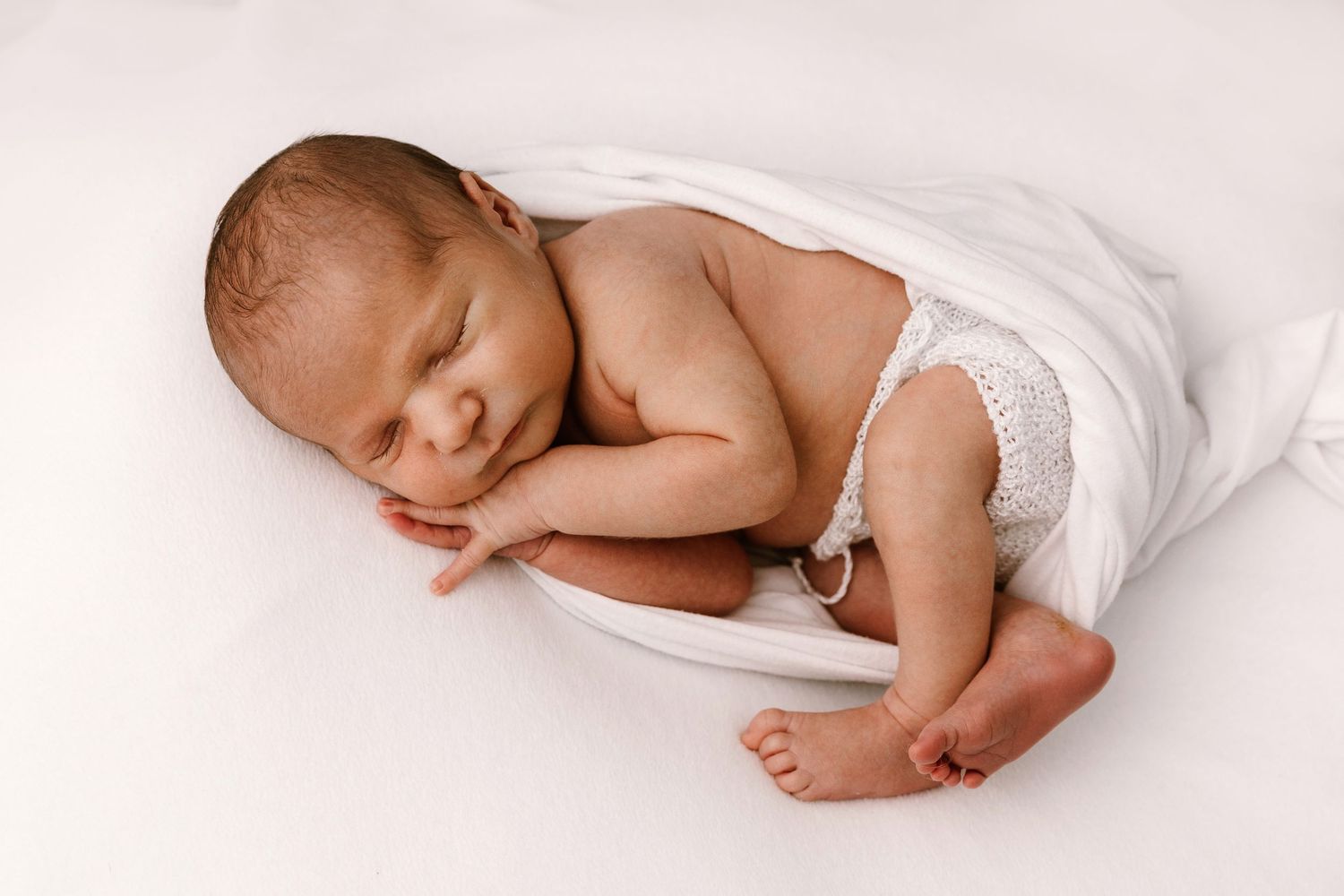 Peaceful newborn baby sleeping wrapped in white swaddle on soft white background in professional photo.