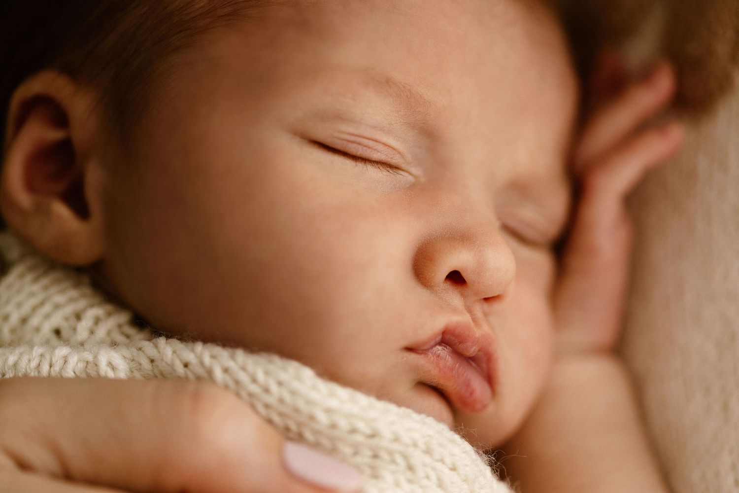 Peaceful newborn baby sleeping with tongue out, wrapped in soft white knit blanket, close-up portrait photography.