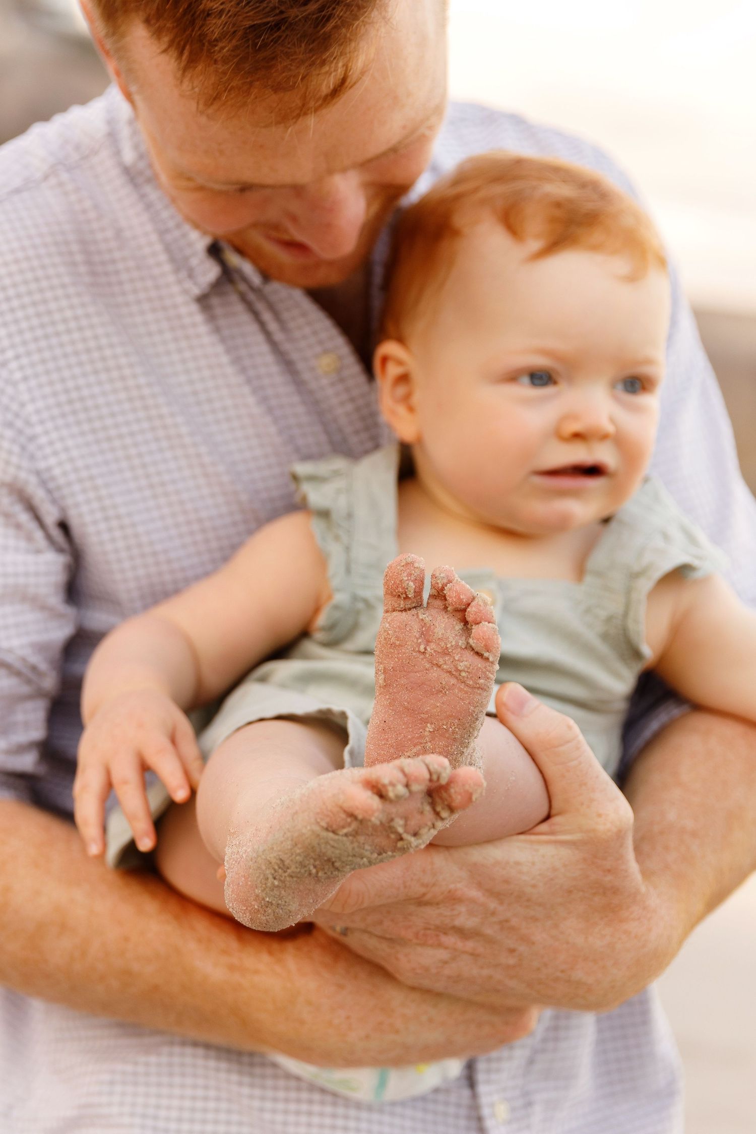 Red-haired father holding baby with sandy feet at the beach.