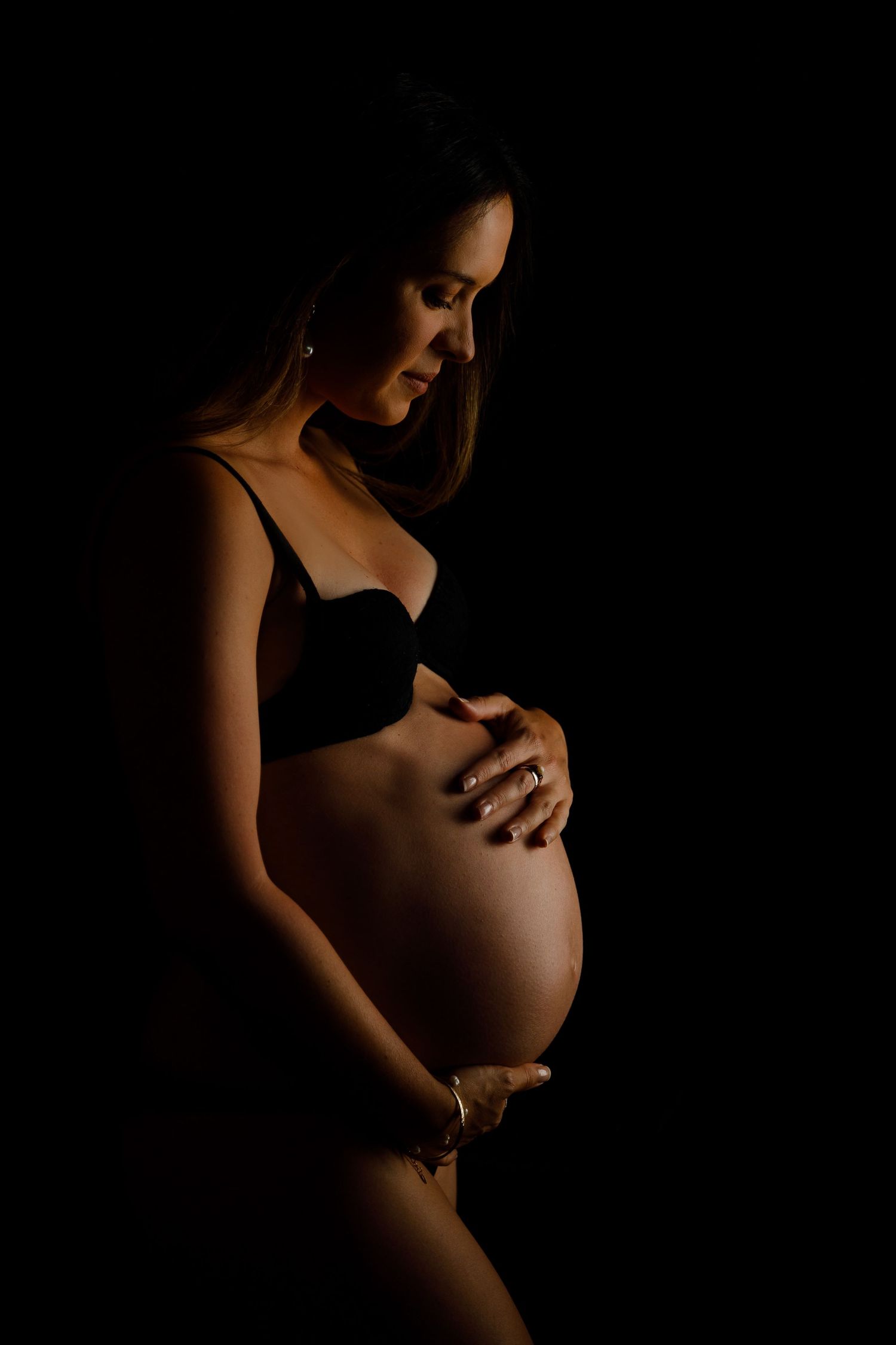 Pregnant woman in black lingerie cradling her baby bump against a dark background in a dramatic studio portrait.